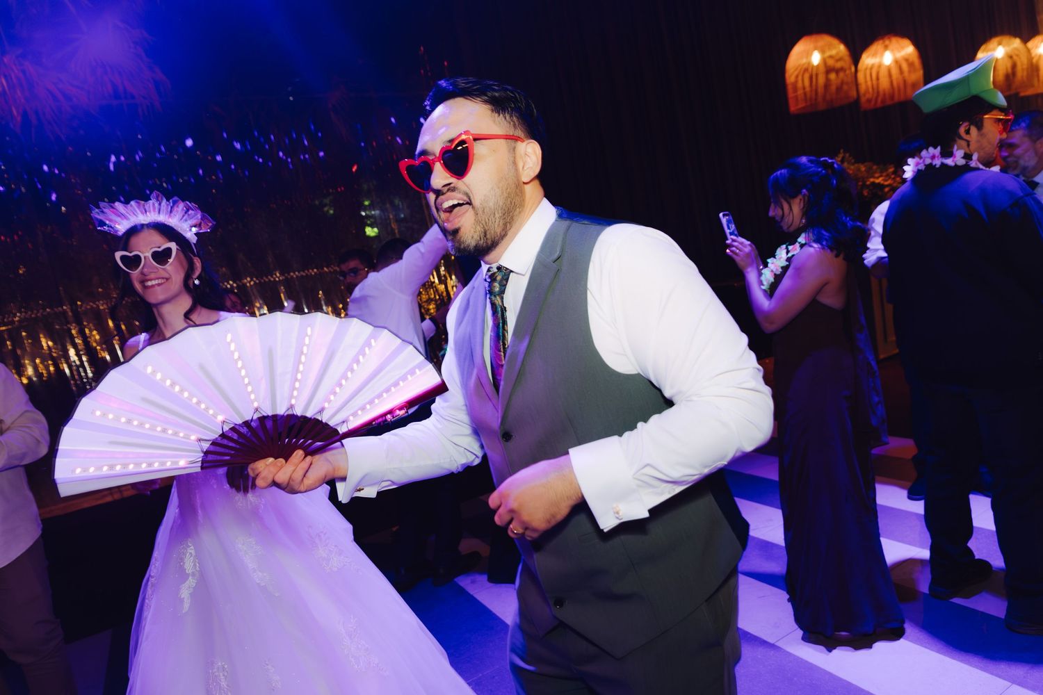 Groom dancing with large white fan while bride wears decorative mask at wedding reception.