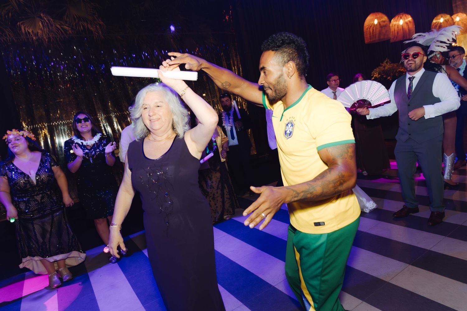 Elderly woman dancing with man in Brazilian soccer jersey at lively wedding reception.