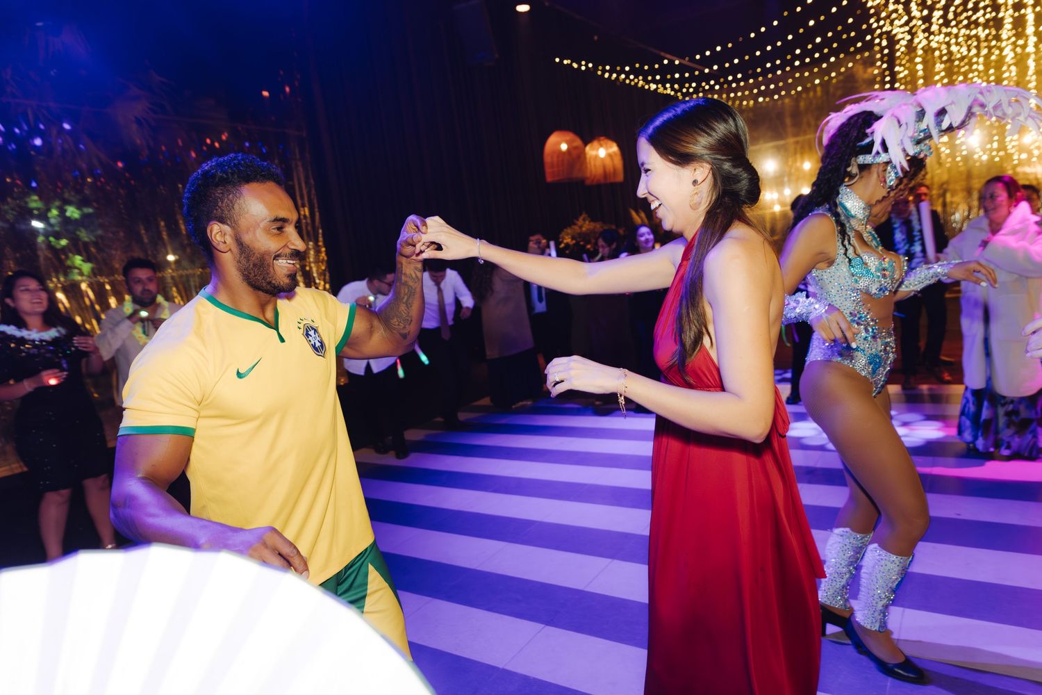 Couple dancing together on a striped dance floor with a samba performer in the background at a festive event.