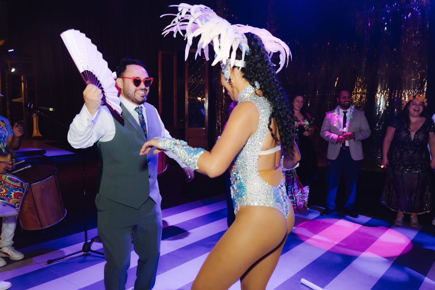 Man in a gray suit dancing with a samba performer wearing a feathered headdress at a lively party.