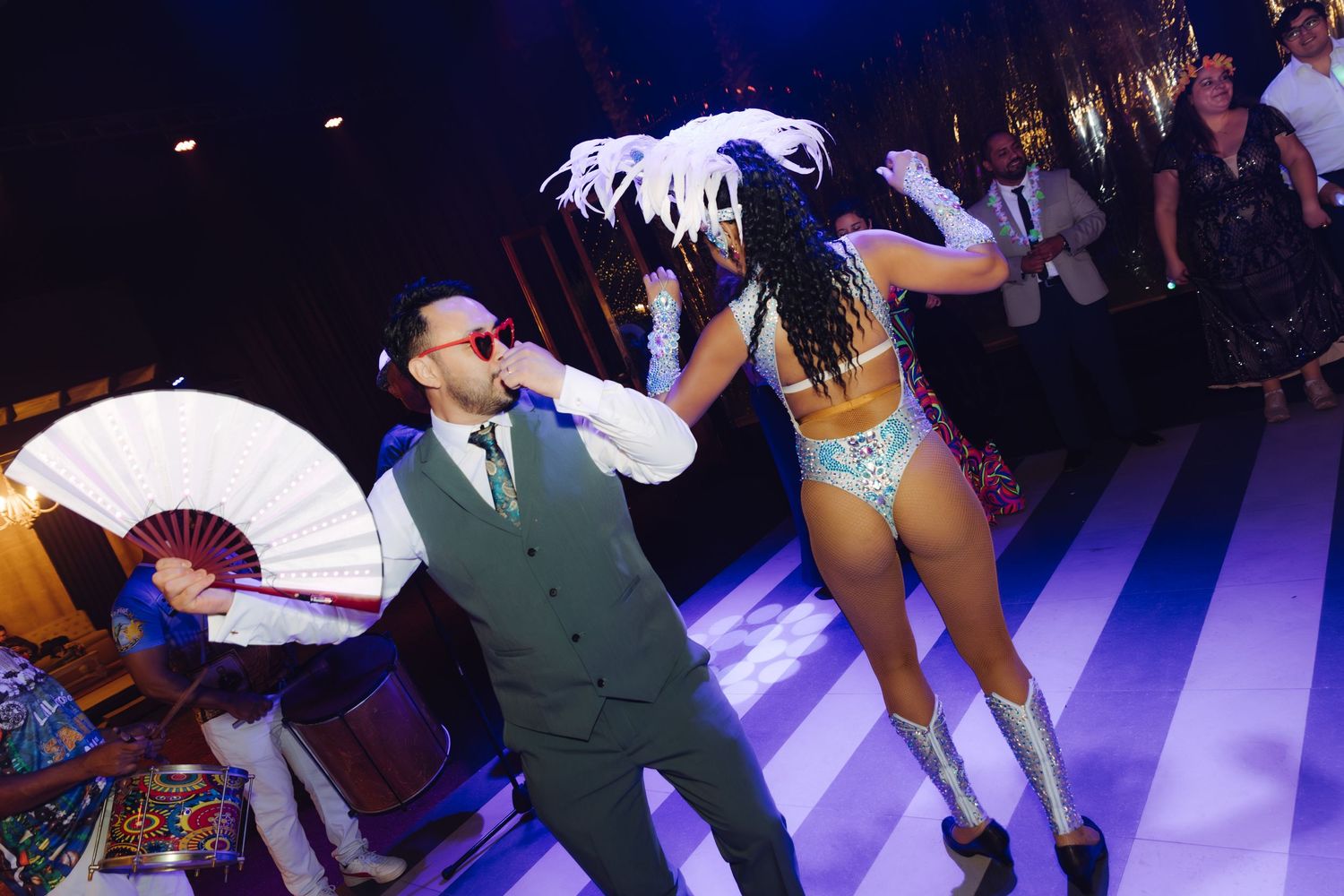 Man in gray suit holding a white fan dancing with a samba performer in a sequined costume at a party.