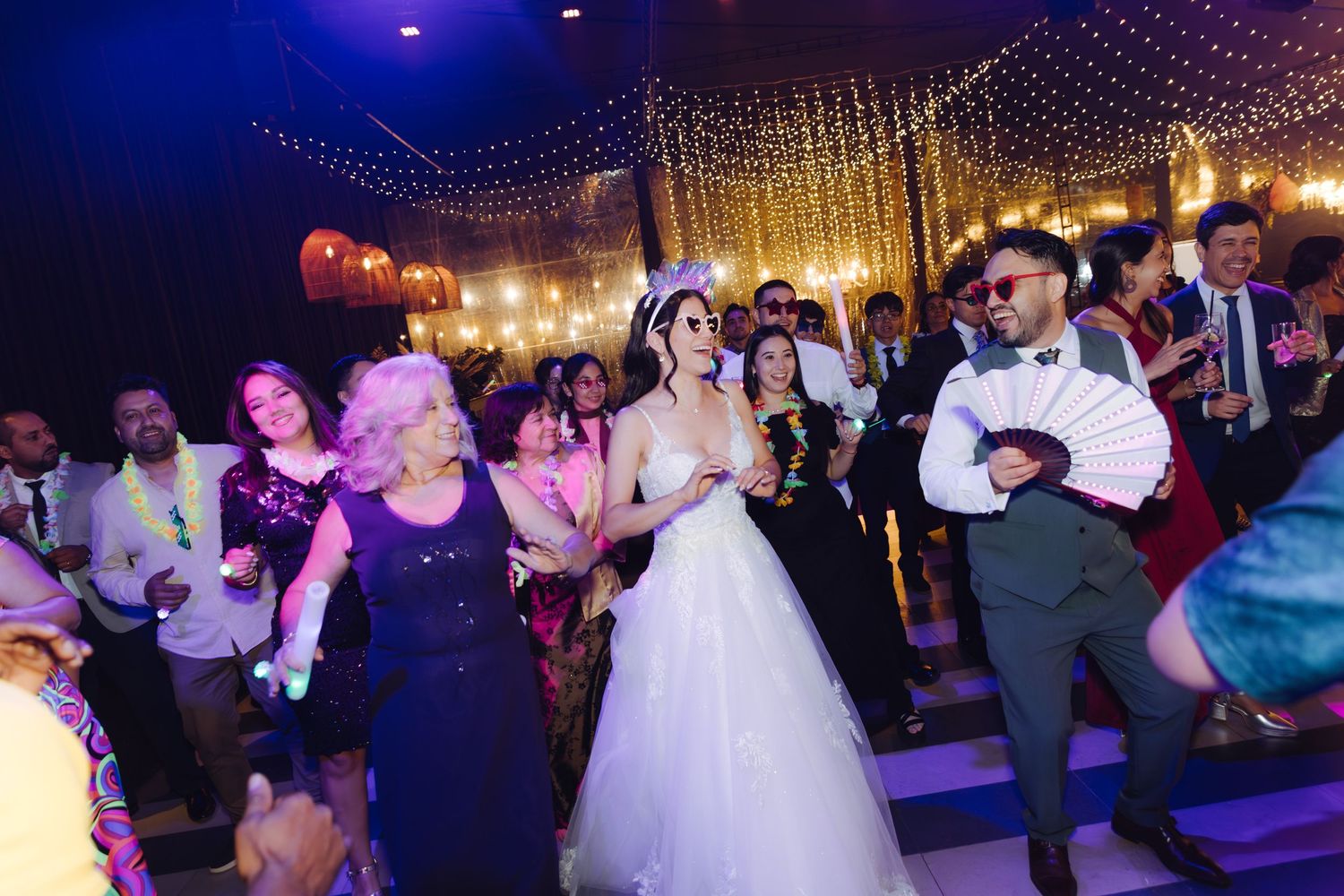 Bride in white gown dancing joyfully with wedding guests under warm string lights at a reception.