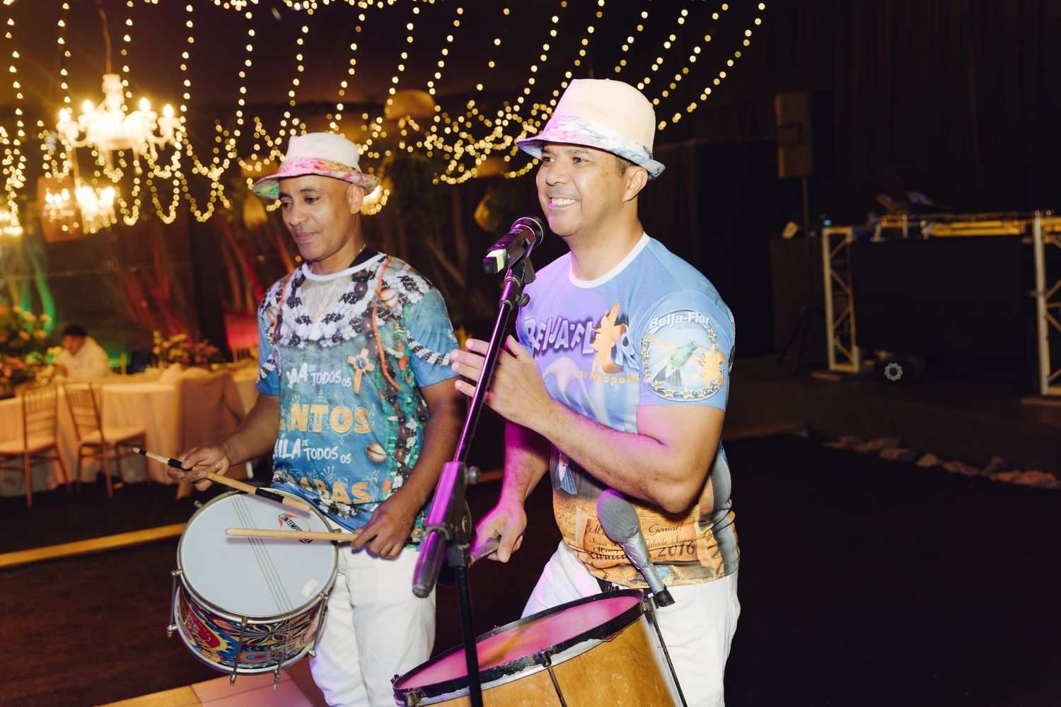 Two musicians in colorful shirts performing with drums and microphone at an outdoor evening event.
