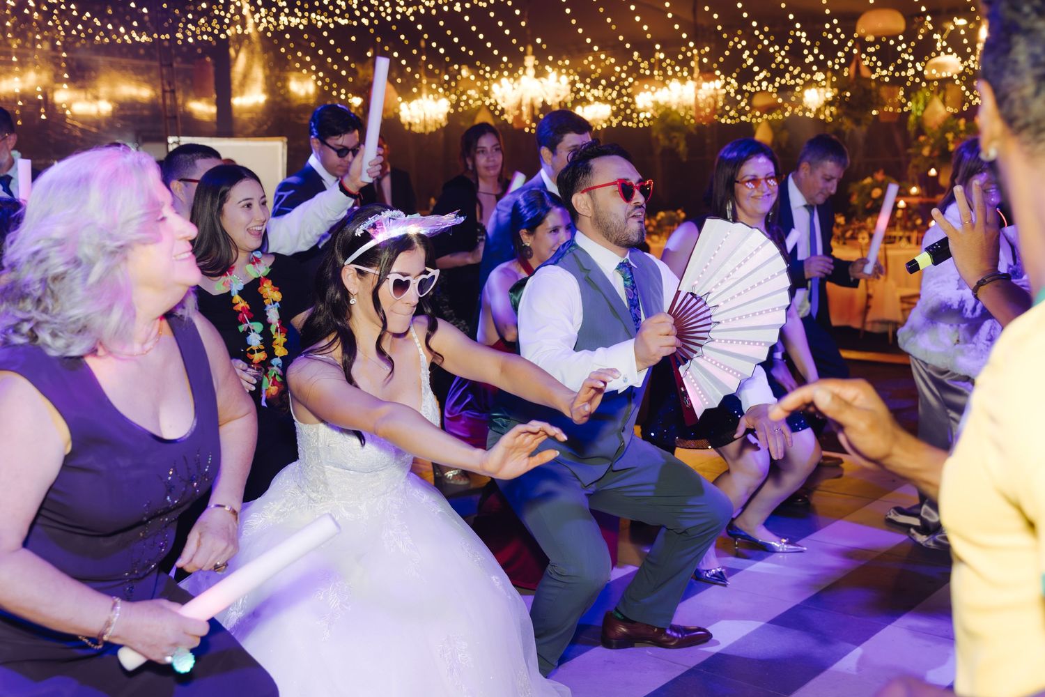 Bride and groom in sunglasses dancing with wedding guests holding glow sticks at a festive reception.