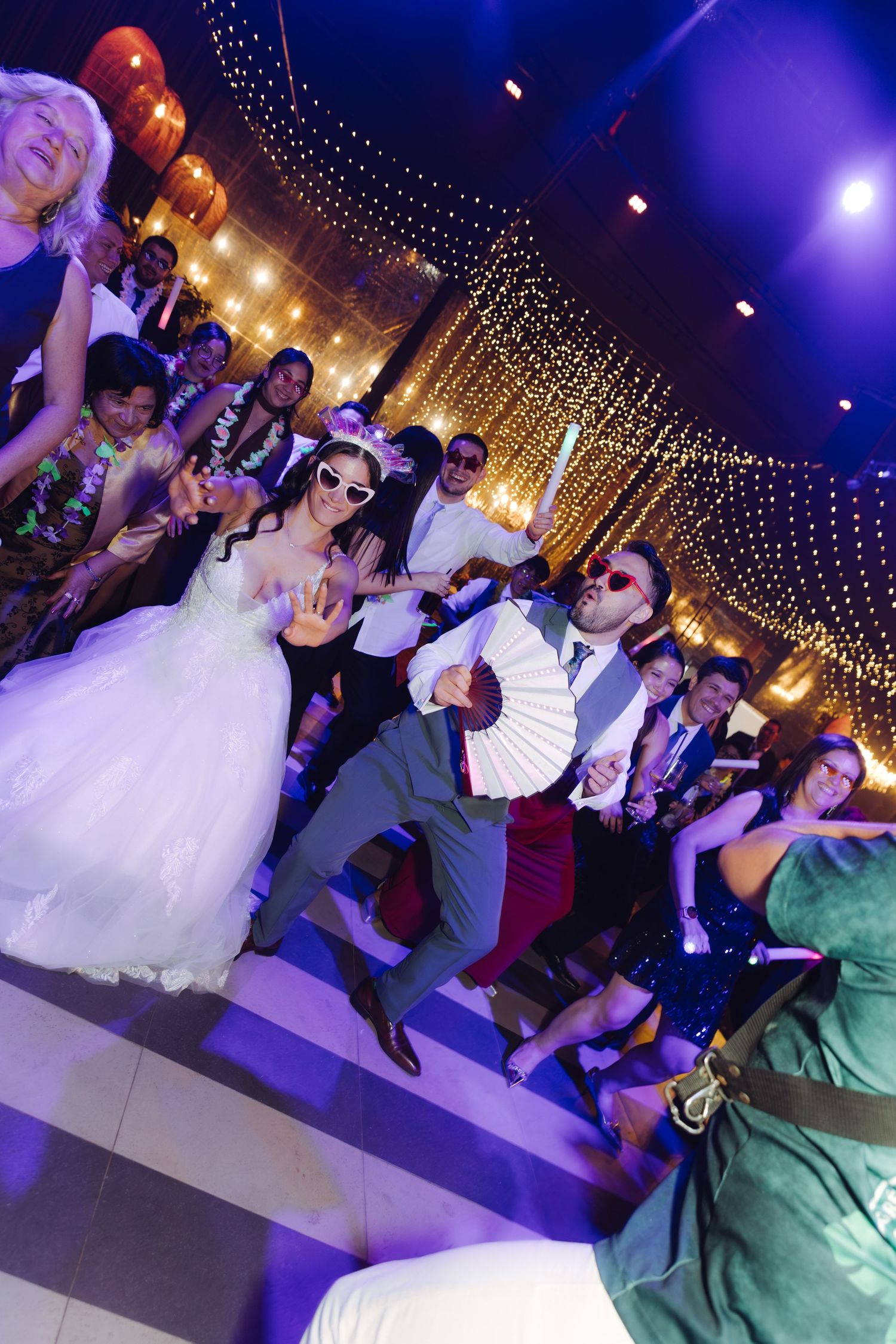Bride and groom wearing sunglasses dancing with wedding guests under twinkling string lights at reception.