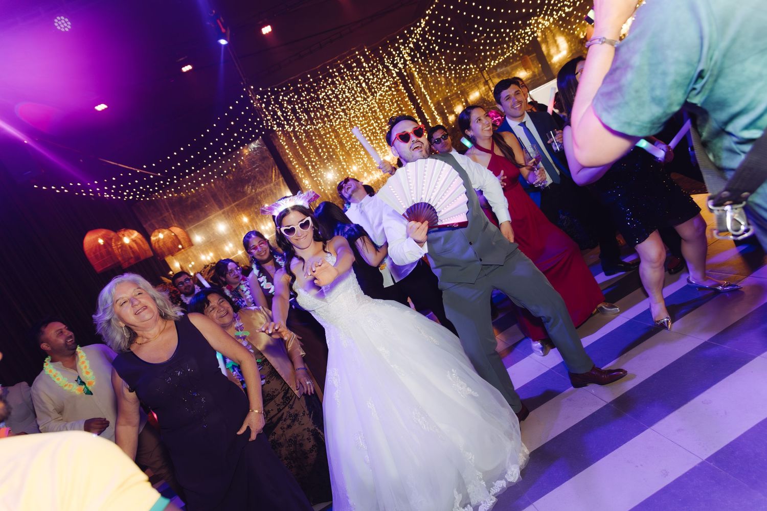 Bride in white gown and groom in gray suit dancing with guests under warm lights at a wedding reception.
