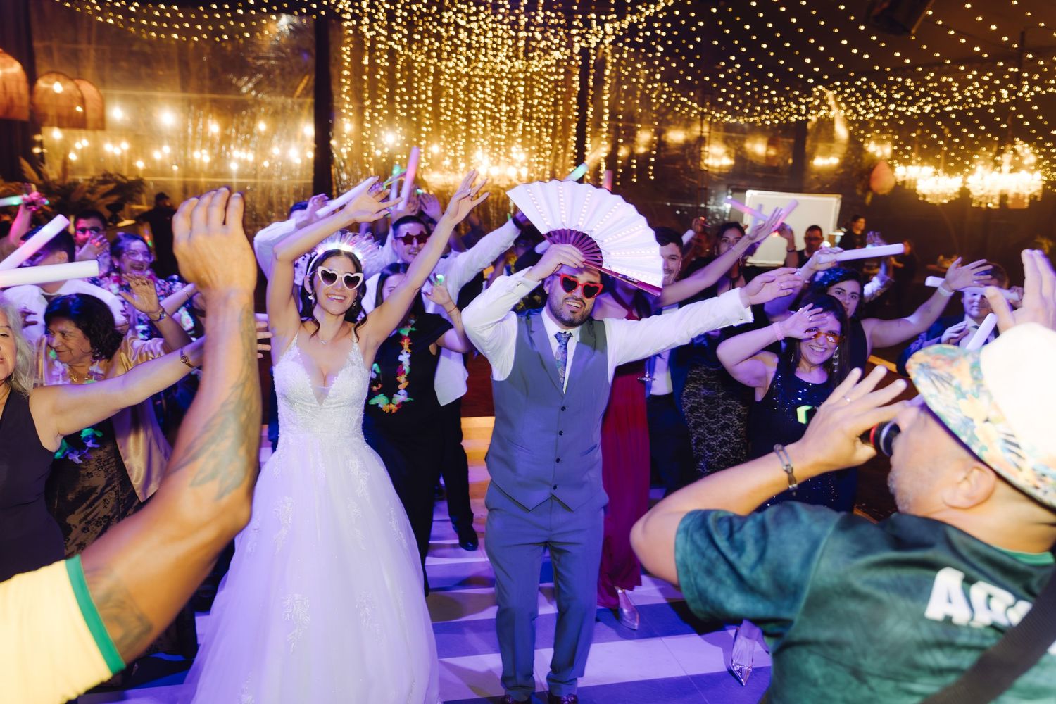Bride and groom dancing with guests at a lively wedding reception under warm string lights.