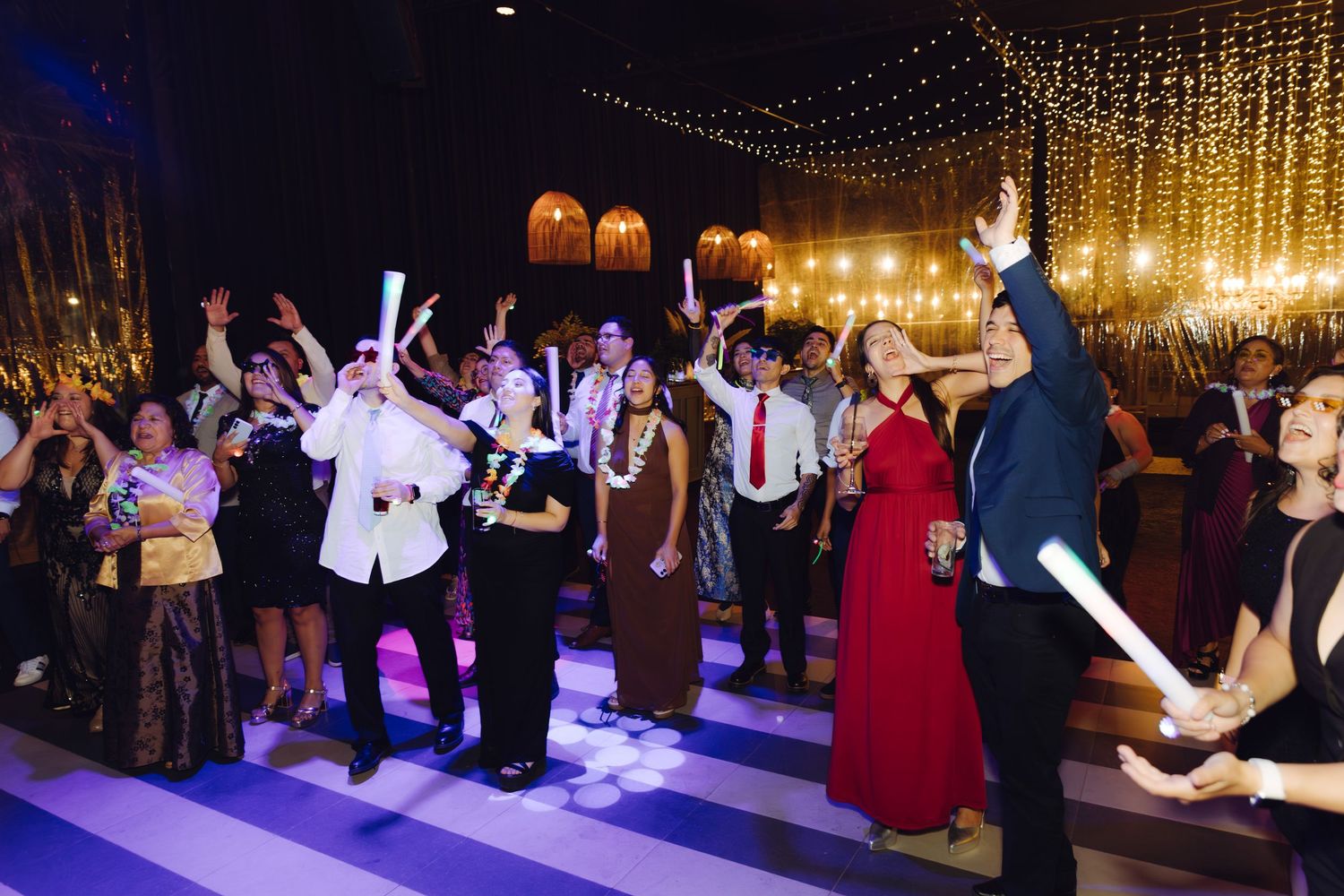 Wedding guests dancing and waving glow sticks on a striped dance floor under twinkling lights.