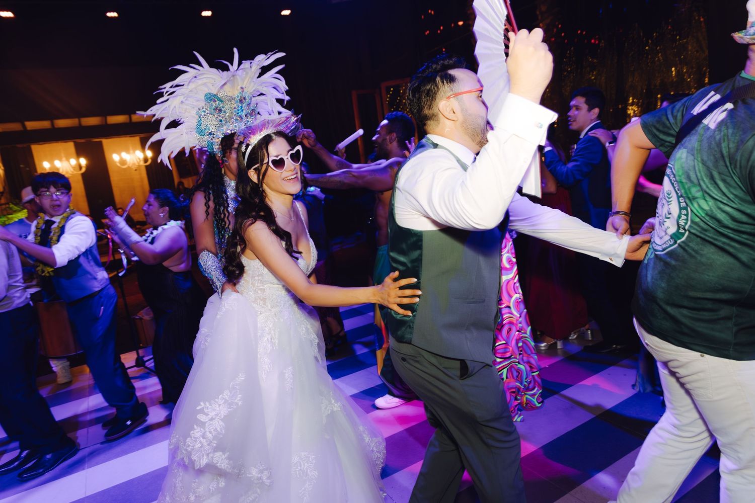 Bride in feathered headpiece and sunglasses dances joyfully with groom at wedding reception.