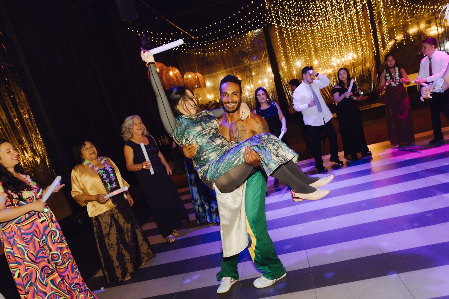 Energetic dancer lifts a guest on a striped dance floor at a vibrant wedding reception.