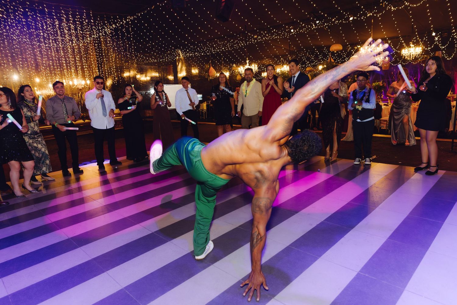 Shirtless acrobatic dancer performs a one-handed handstand on a striped wedding reception dance floor.