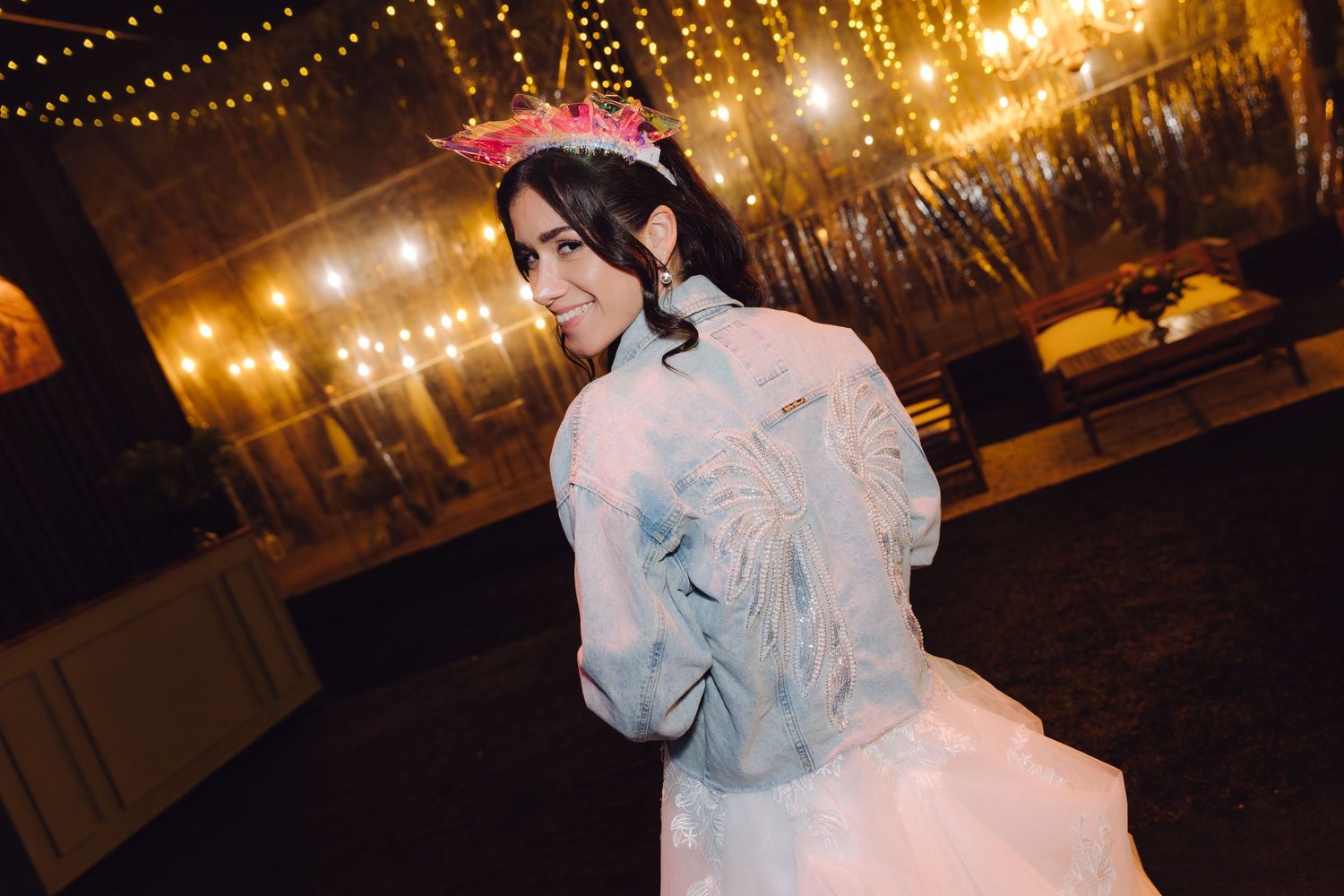 Bride in white gown and floral headpiece smiles outdoors at night with bokeh lights behind her.