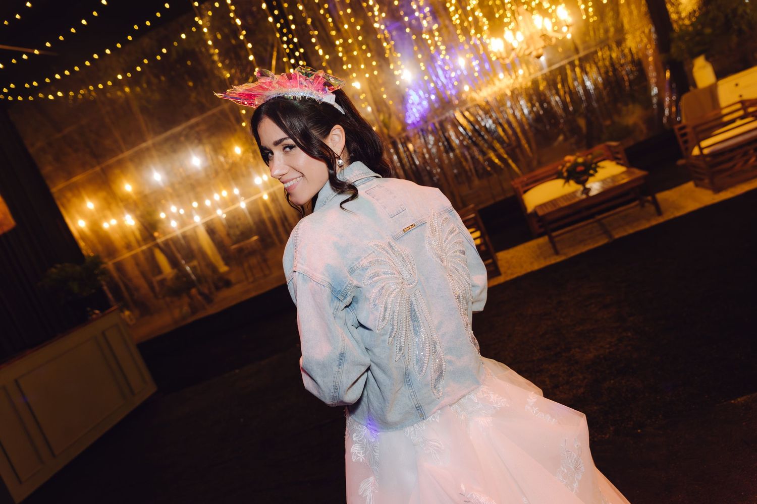 Bride in white wedding gown and floral crown walks outdoors at night under warm string lights.