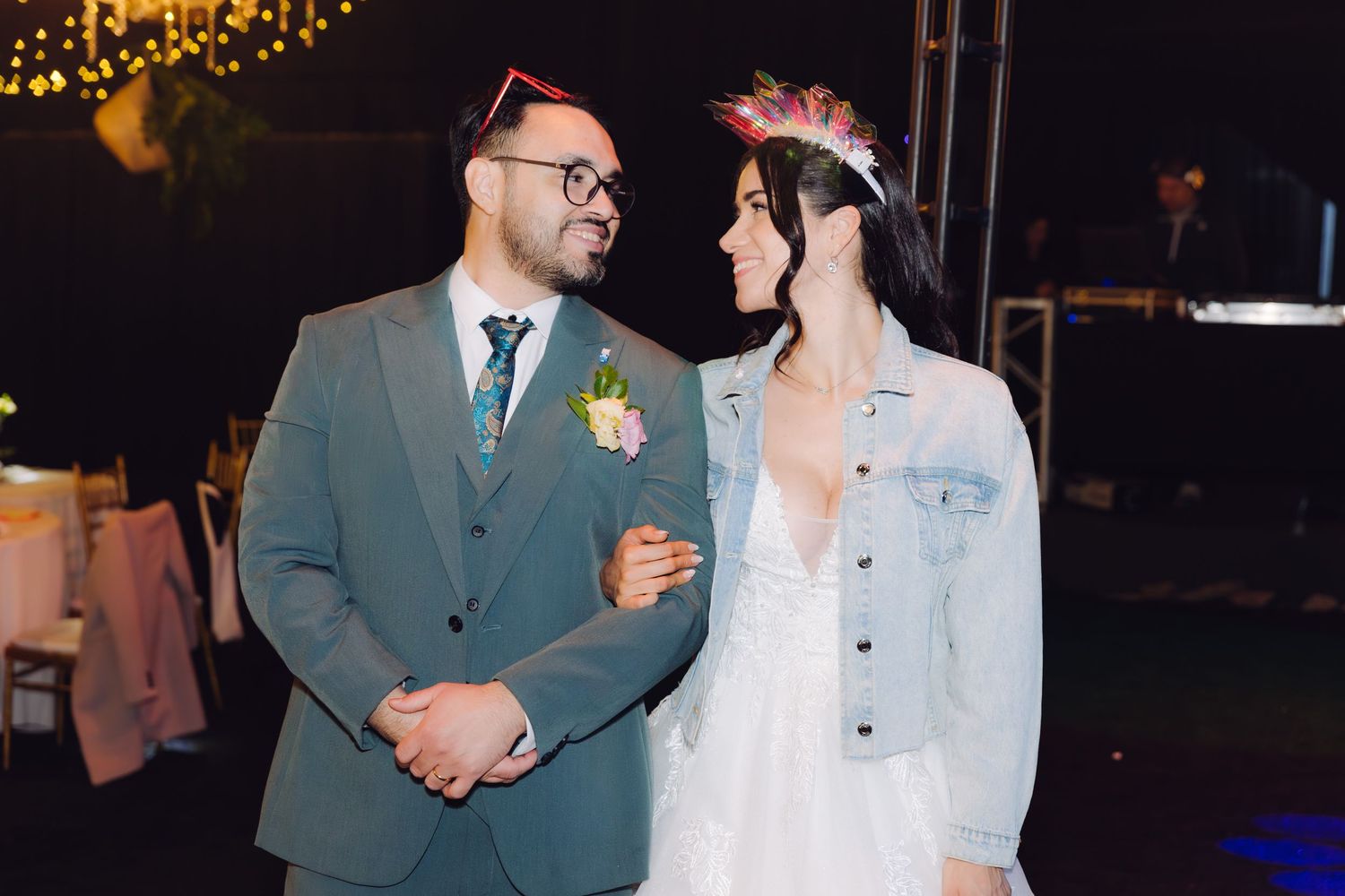 Bride in denim jacket and groom in gray suit smiling at each other at outdoor wedding reception at night.