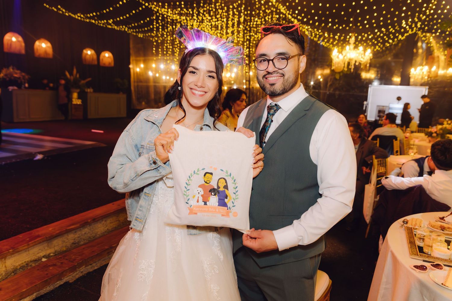 Smiling bride and groom holding illustrated cartoon portrait at festive wedding reception.