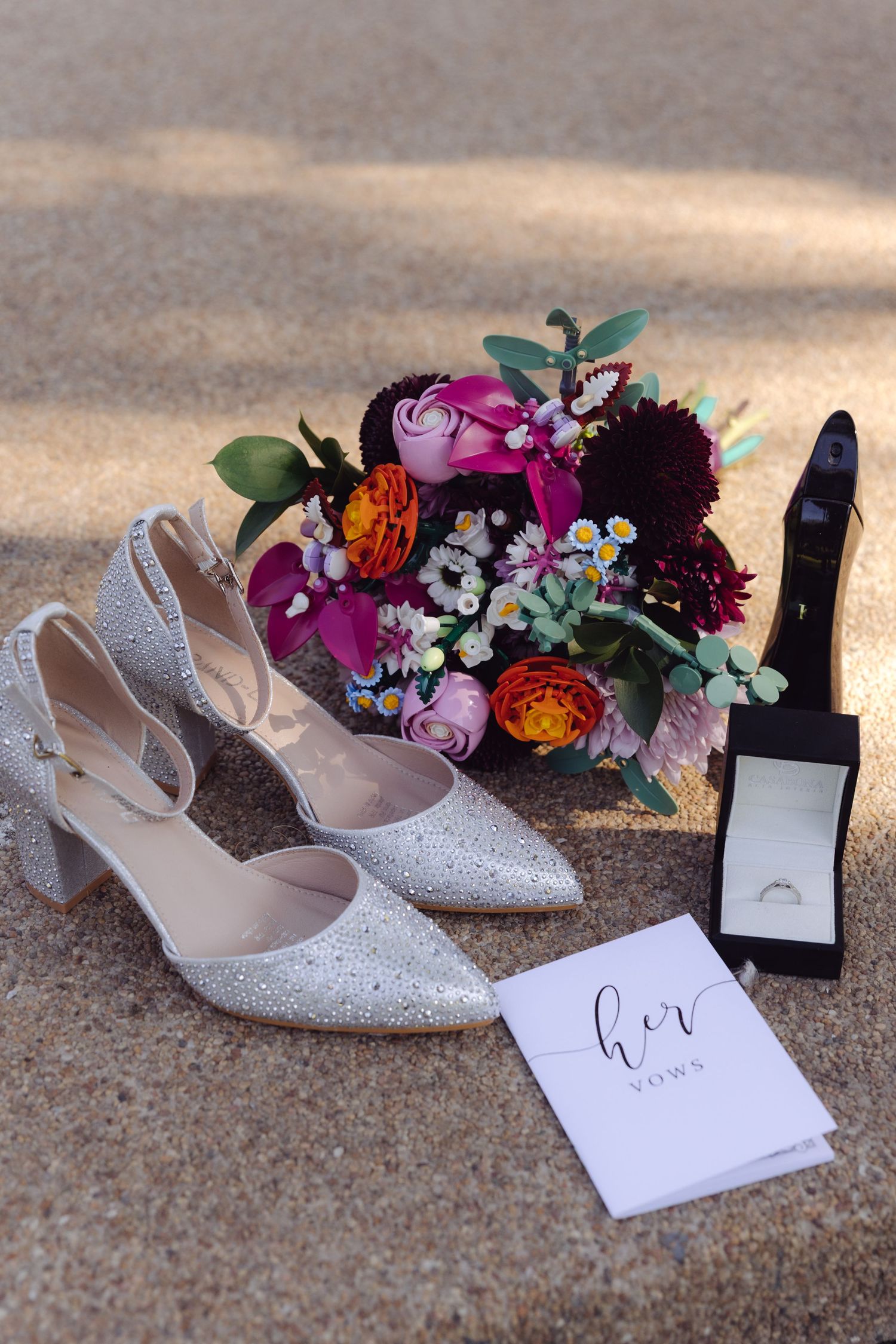 Wedding flat lay featuring glittery heels, colorful bridal bouquet, ring box, and her vows booklet on sandy surface.