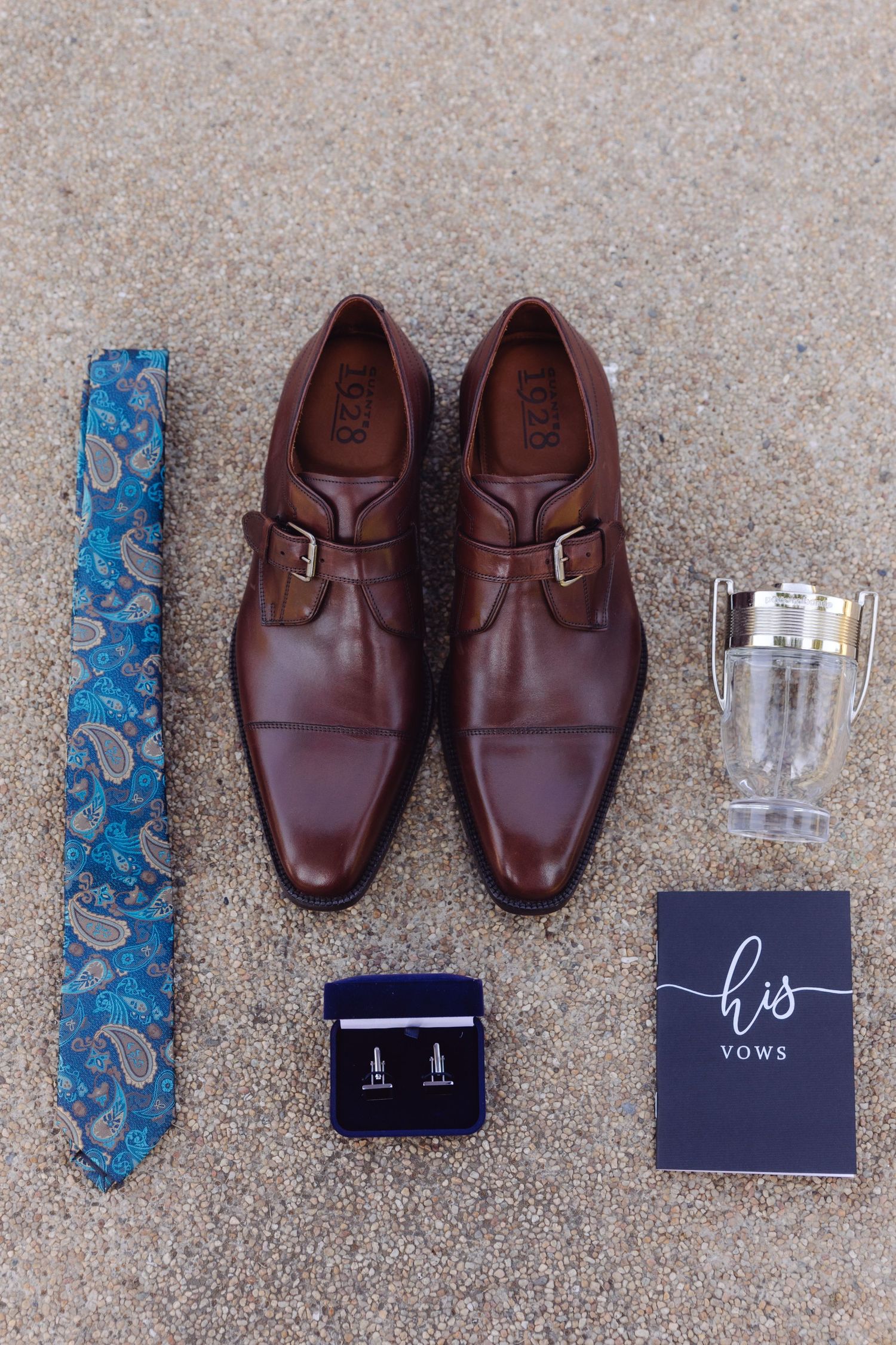 Groom wedding flat lay with brown monk strap shoes, blue paisley tie, cufflinks, cologne, and his vows booklet.