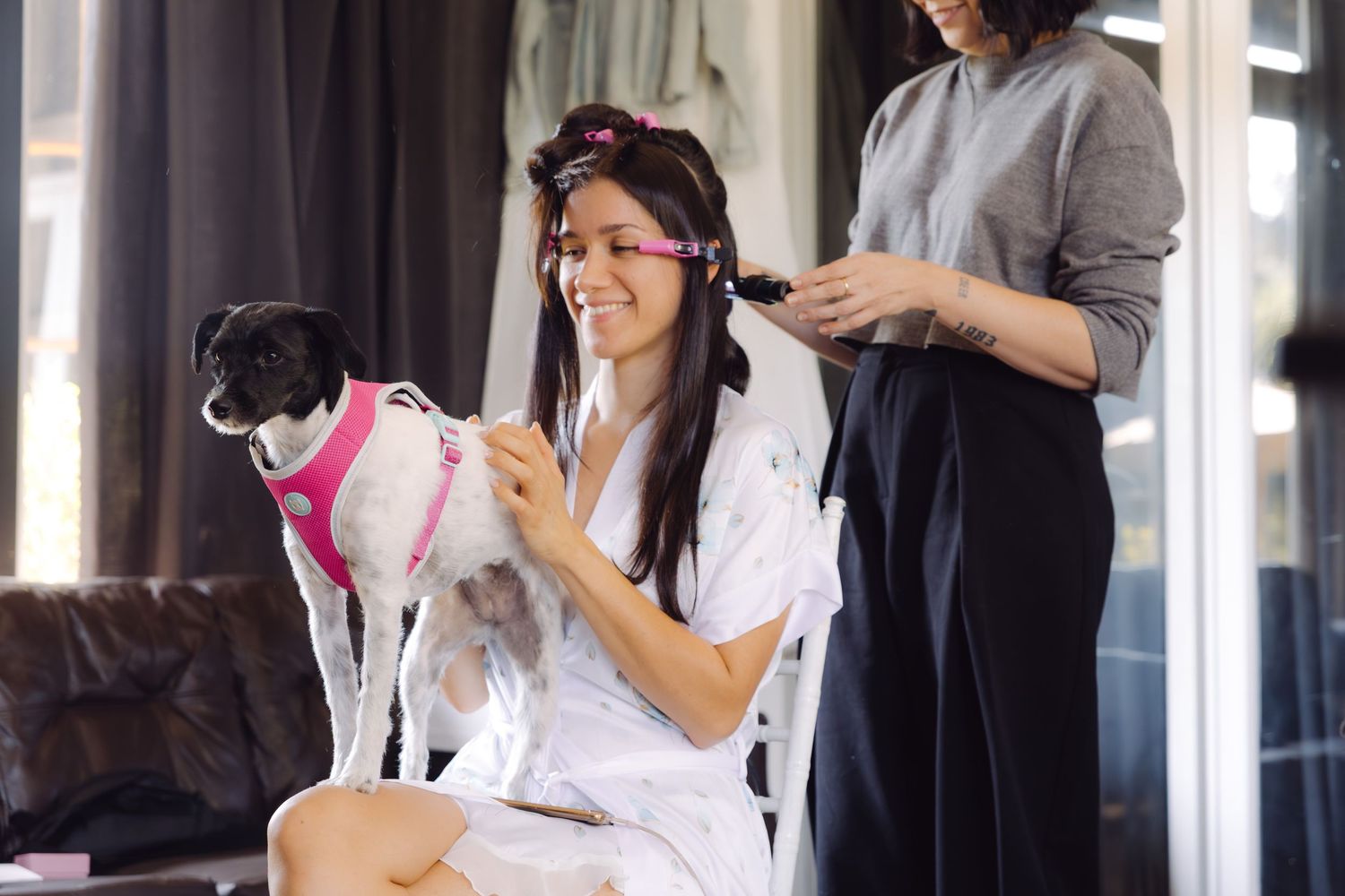 Smiling bride in white robe with hair rollers holds a dog in pink harness while getting hair styled.