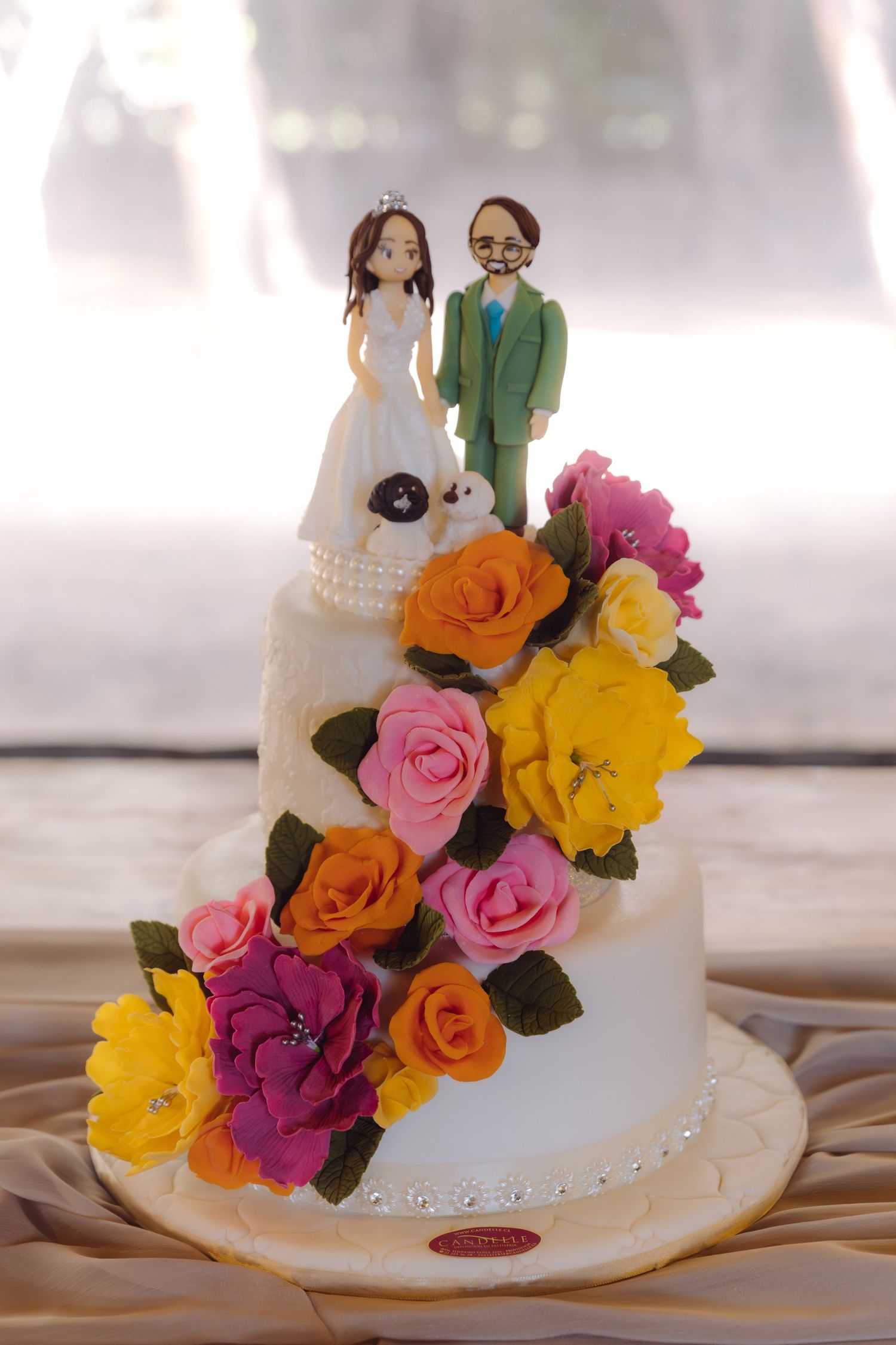 Two-tier white wedding cake topped with custom bride and groom figurines surrounded by vibrant colorful sugar flowers.