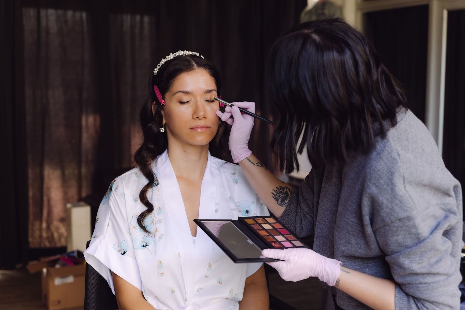 Bride in white robe getting professional makeup applied by artist holding eyeshadow palette.