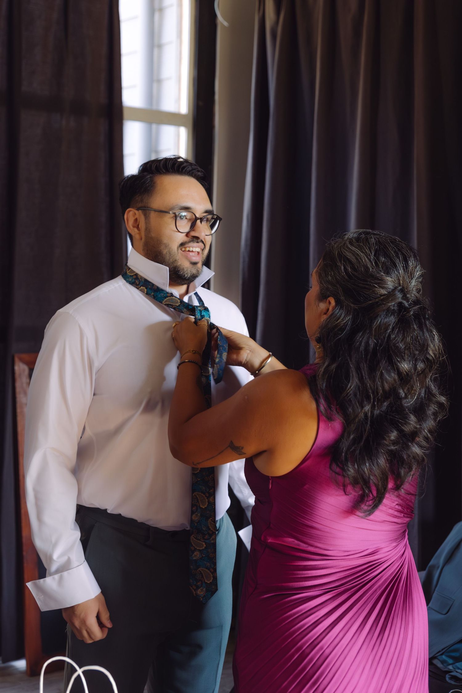 Groom in white shirt smiling as woman in magenta dress helps adjust his floral necktie.