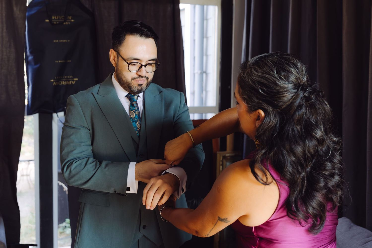 Groom in teal three-piece suit having cufflinks fastened by woman in magenta dress.