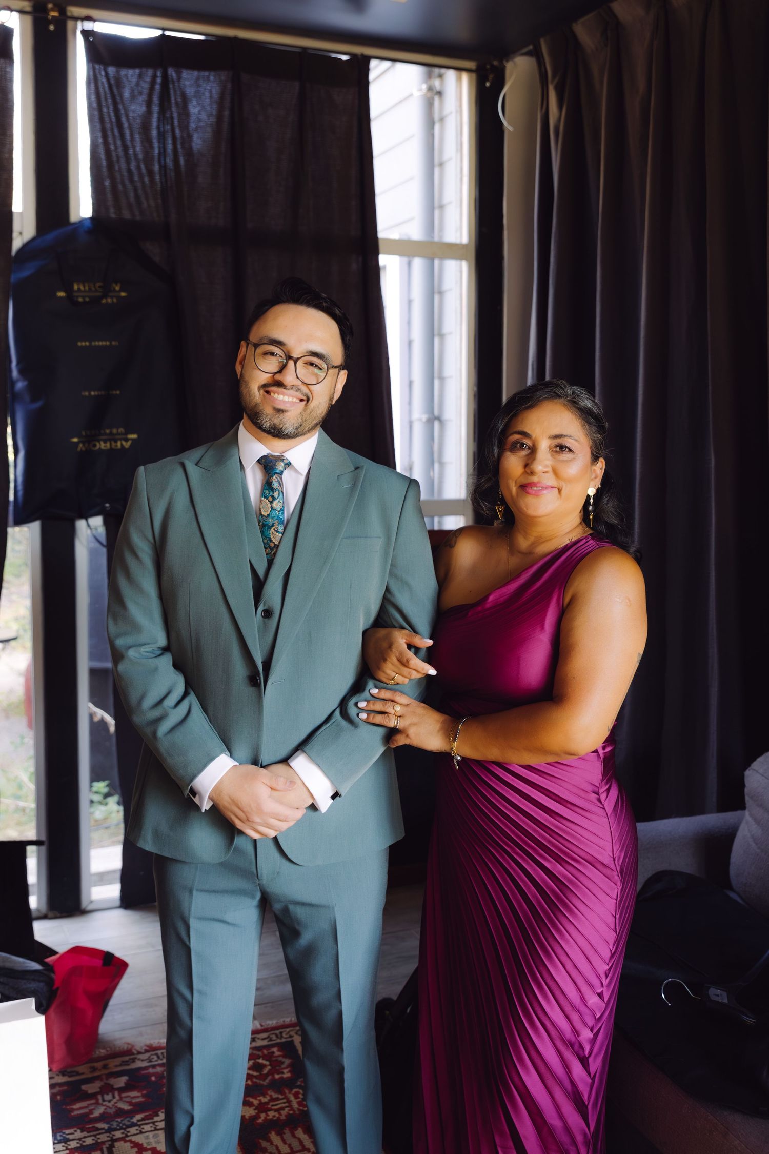 Groom in teal three-piece suit posing with smiling woman in magenta gown before wedding ceremony.