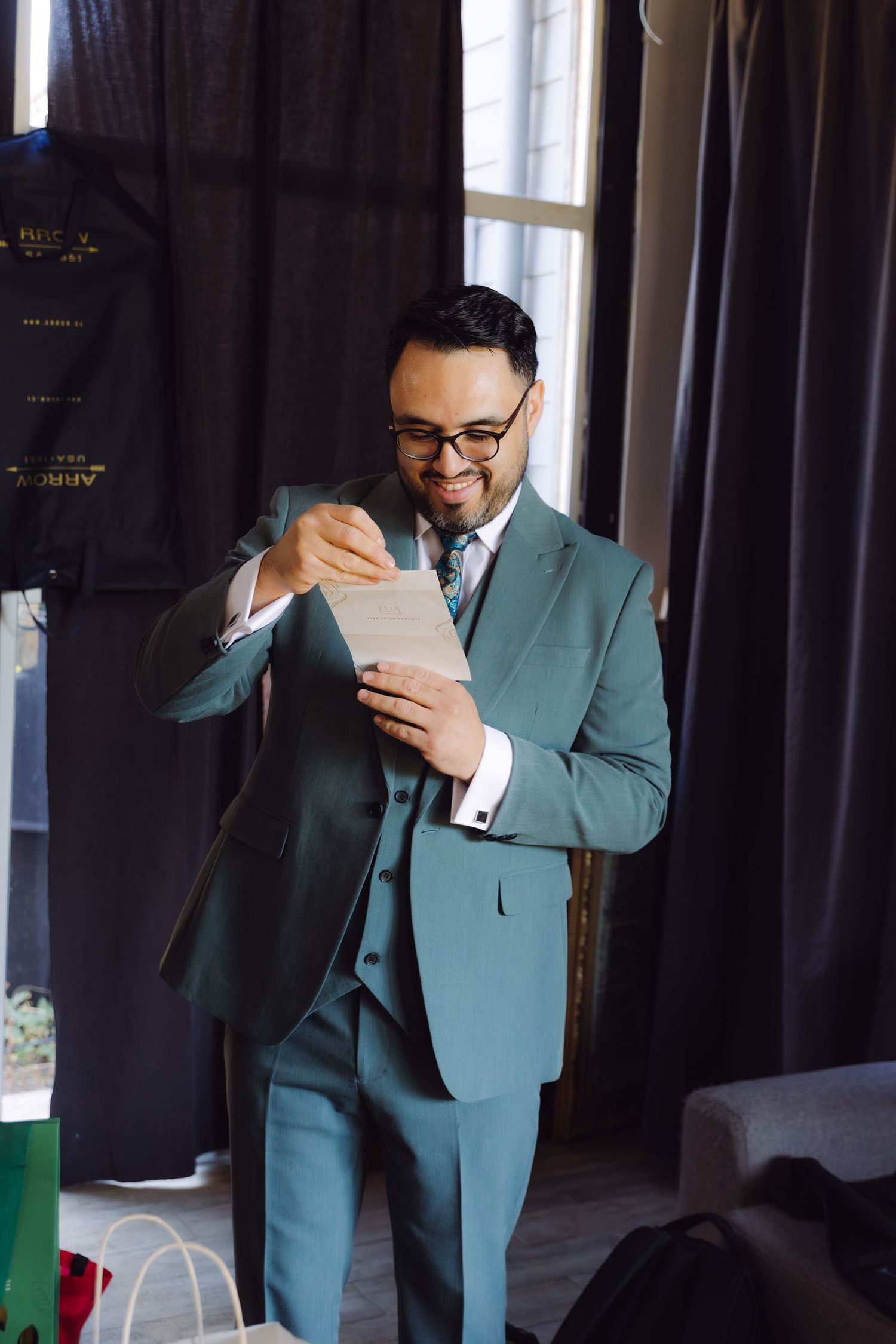 Handsome groom in teal three-piece suit smiling while reading a heartfelt letter before his wedding.