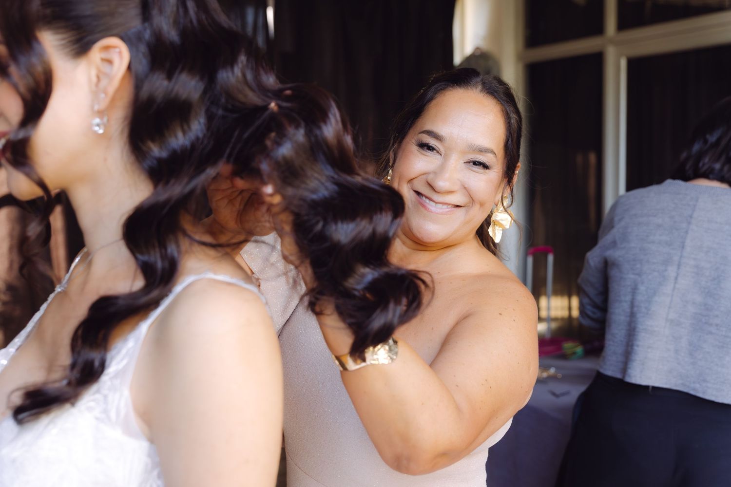 Bridesmaid smiling while styling the bride's dark curly hair during wedding preparations.