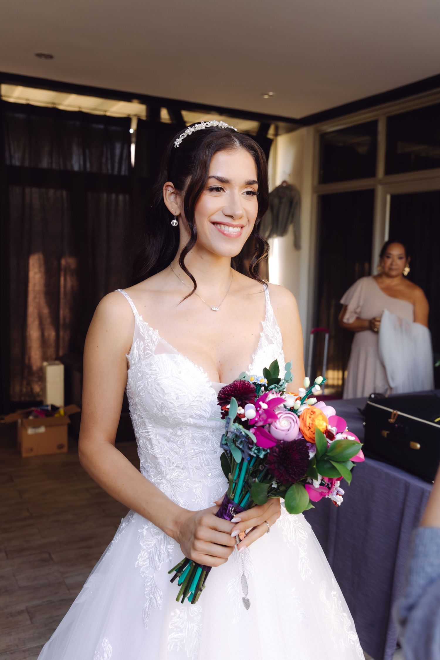 Smiling bride in white lace gown holding colorful bridal bouquet with crystal tiara in bridal suite.
