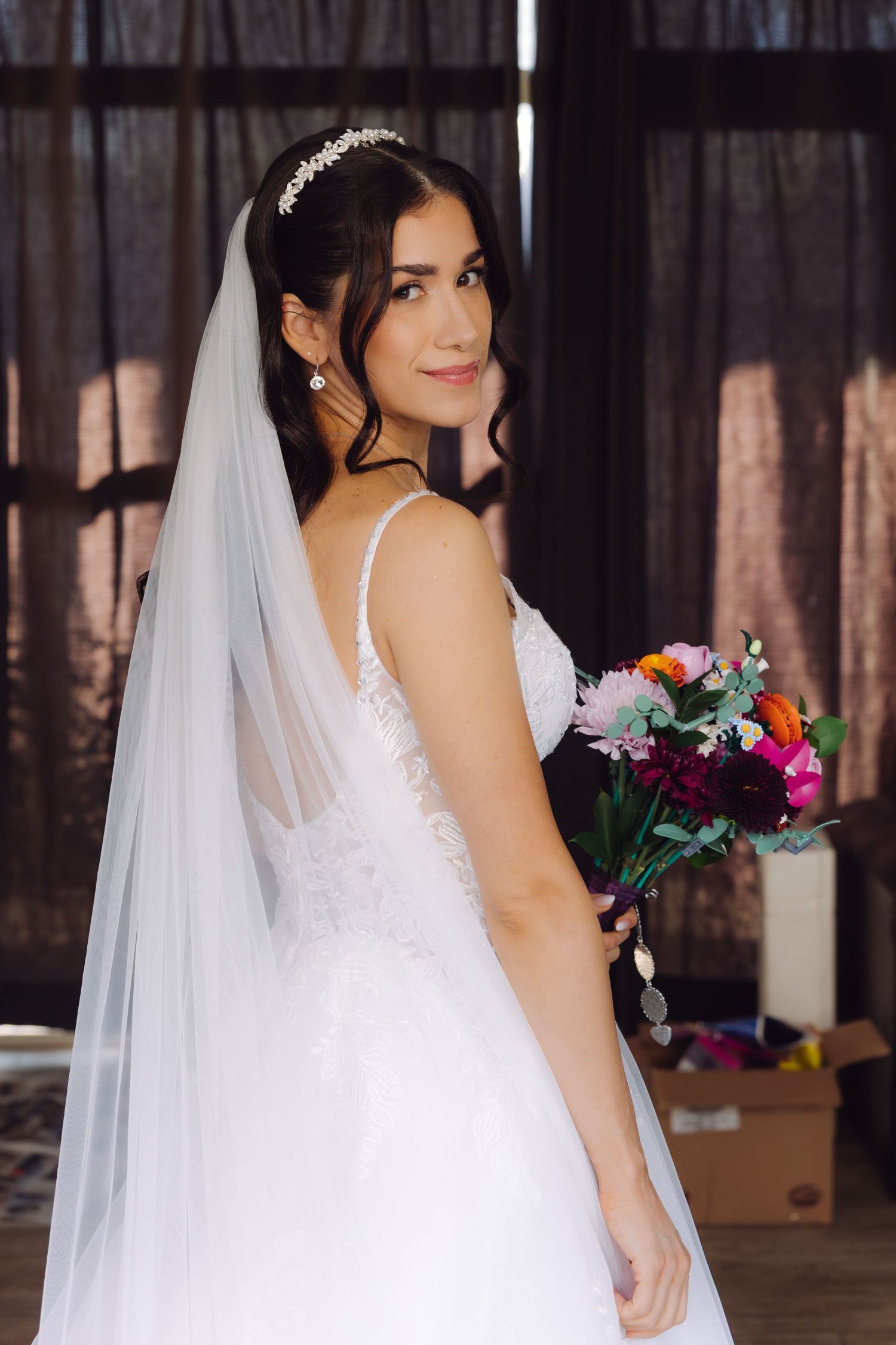 Elegant bride in white lace gown with veil and tiara holding colorful bouquet, looking over shoulder.