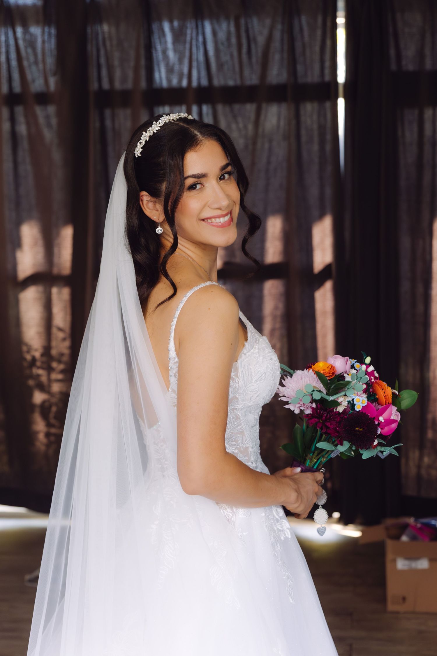 Bride in white wedding dress and long veil holding colorful bouquet while smiling over her shoulder.