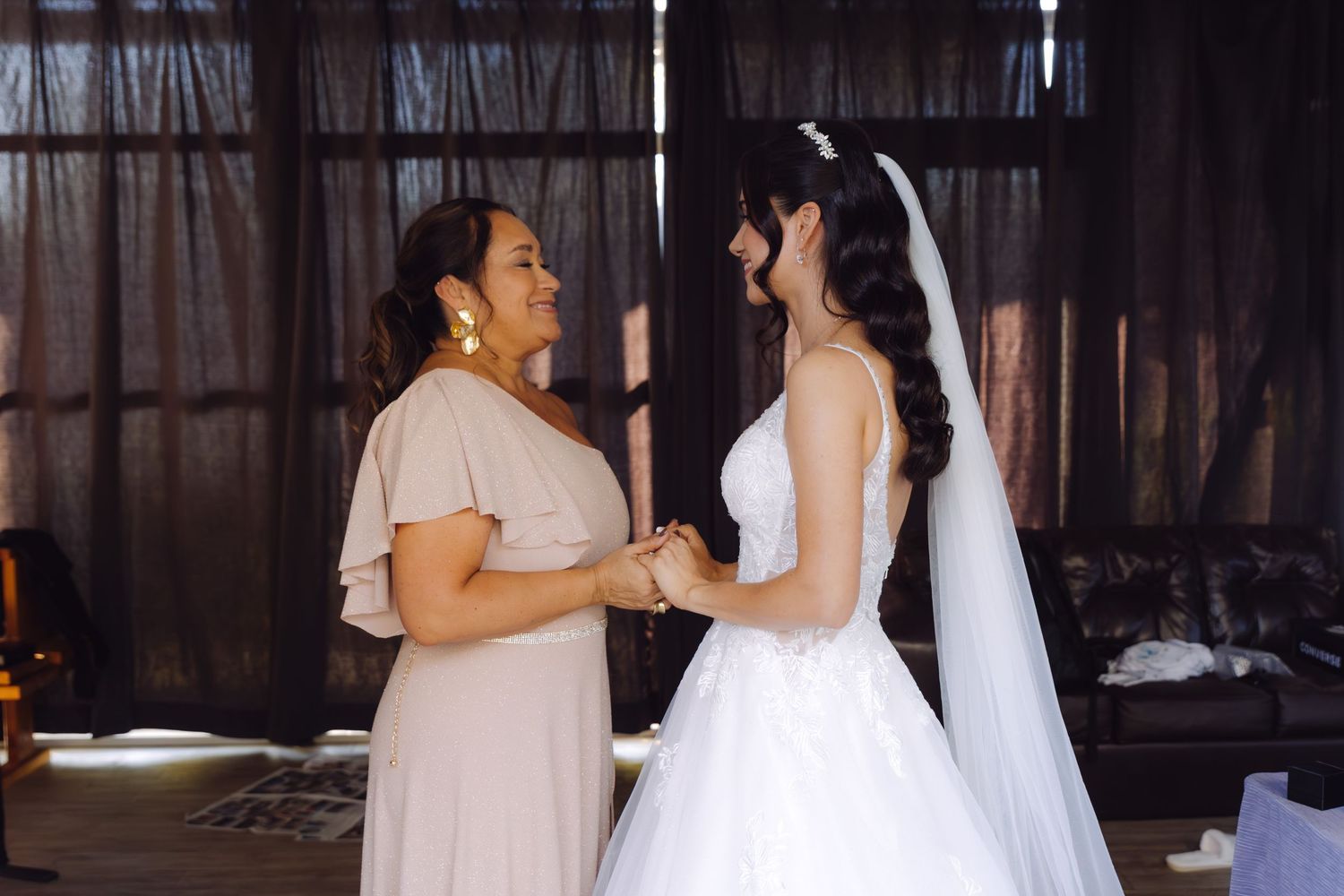 Emotional moment between bride in white gown with veil and woman in beige dress holding hands.