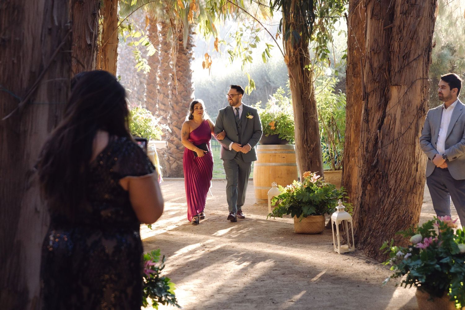 Groom in gray suit walks with bridesmaid in red dress through tree-lined outdoor wedding venue.