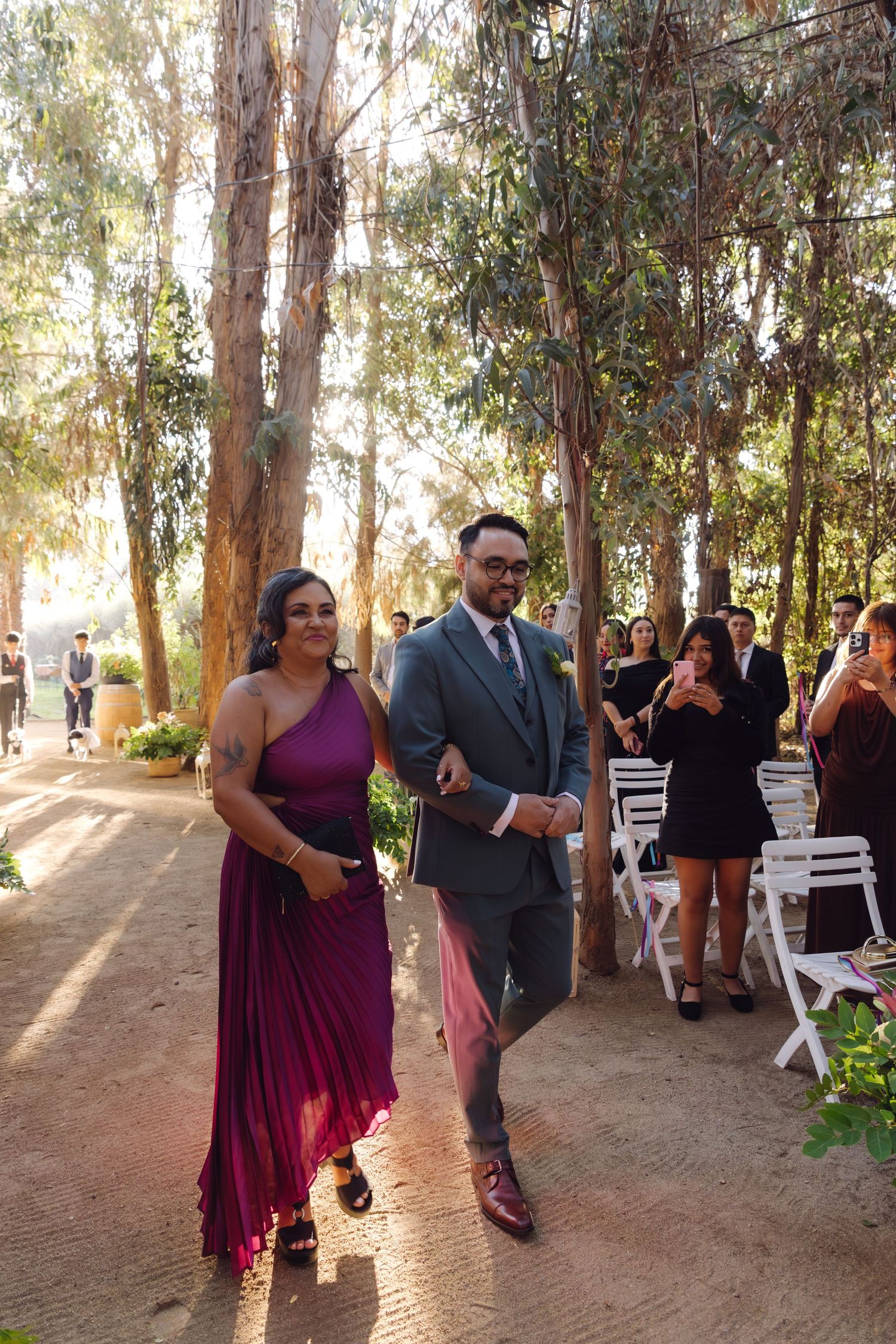 Groom in gray suit escorts bridesmaid in purple dress down outdoor wedding ceremony aisle.