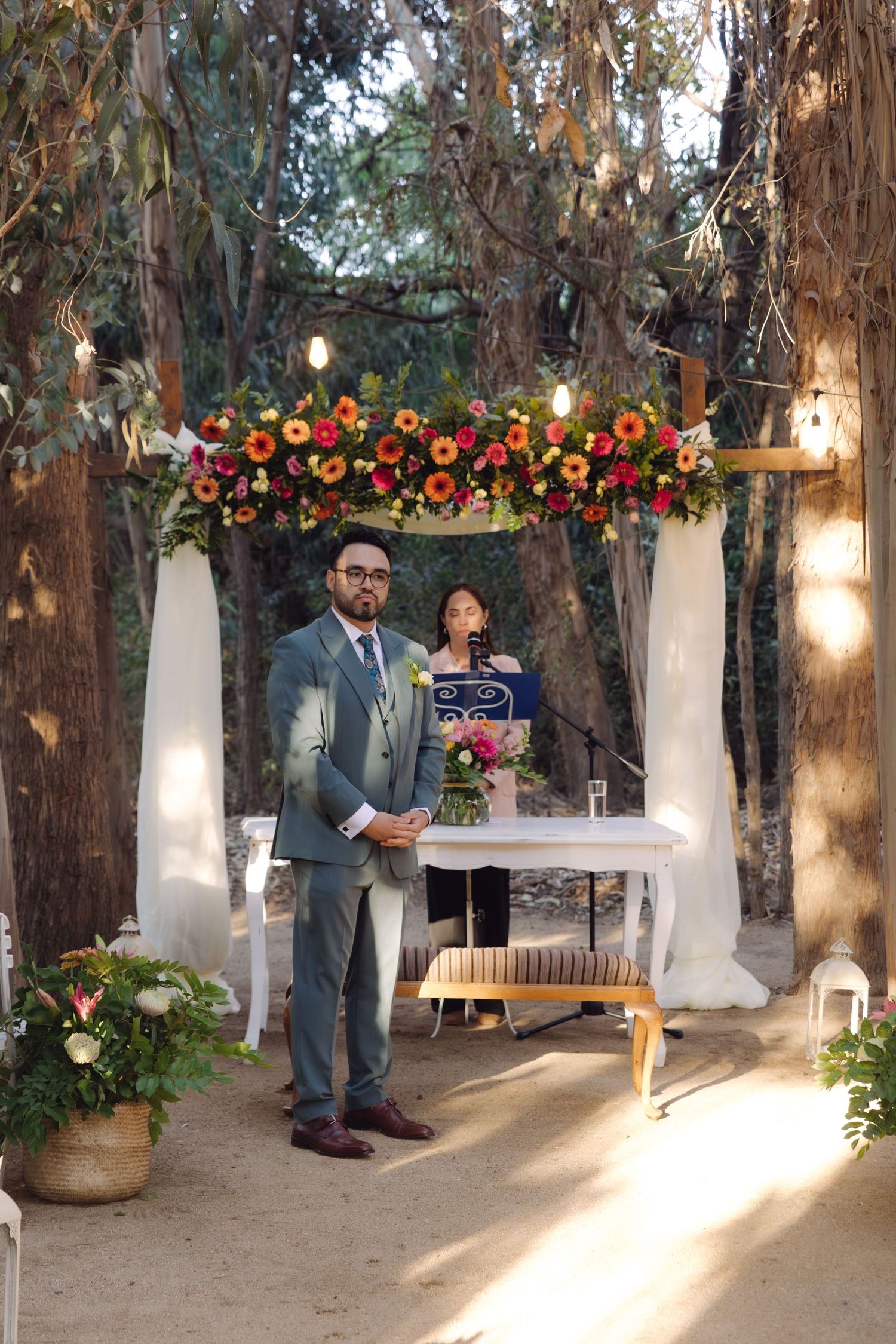 Groom in gray suit stands at floral arch altar with officiant during outdoor woodland wedding.