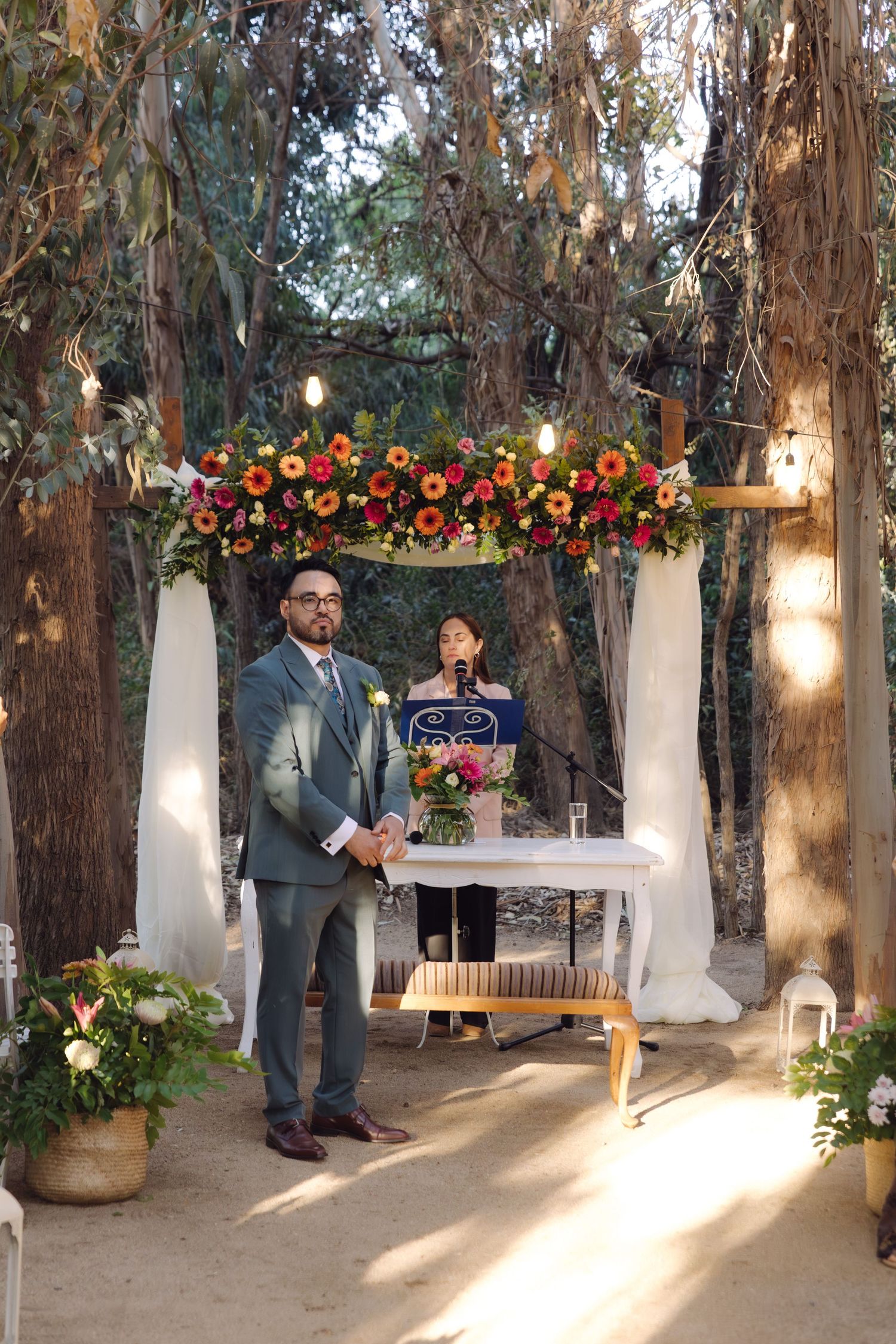Groom in gray suit stands beneath colorful floral wedding arch as officiant prepares ceremony.