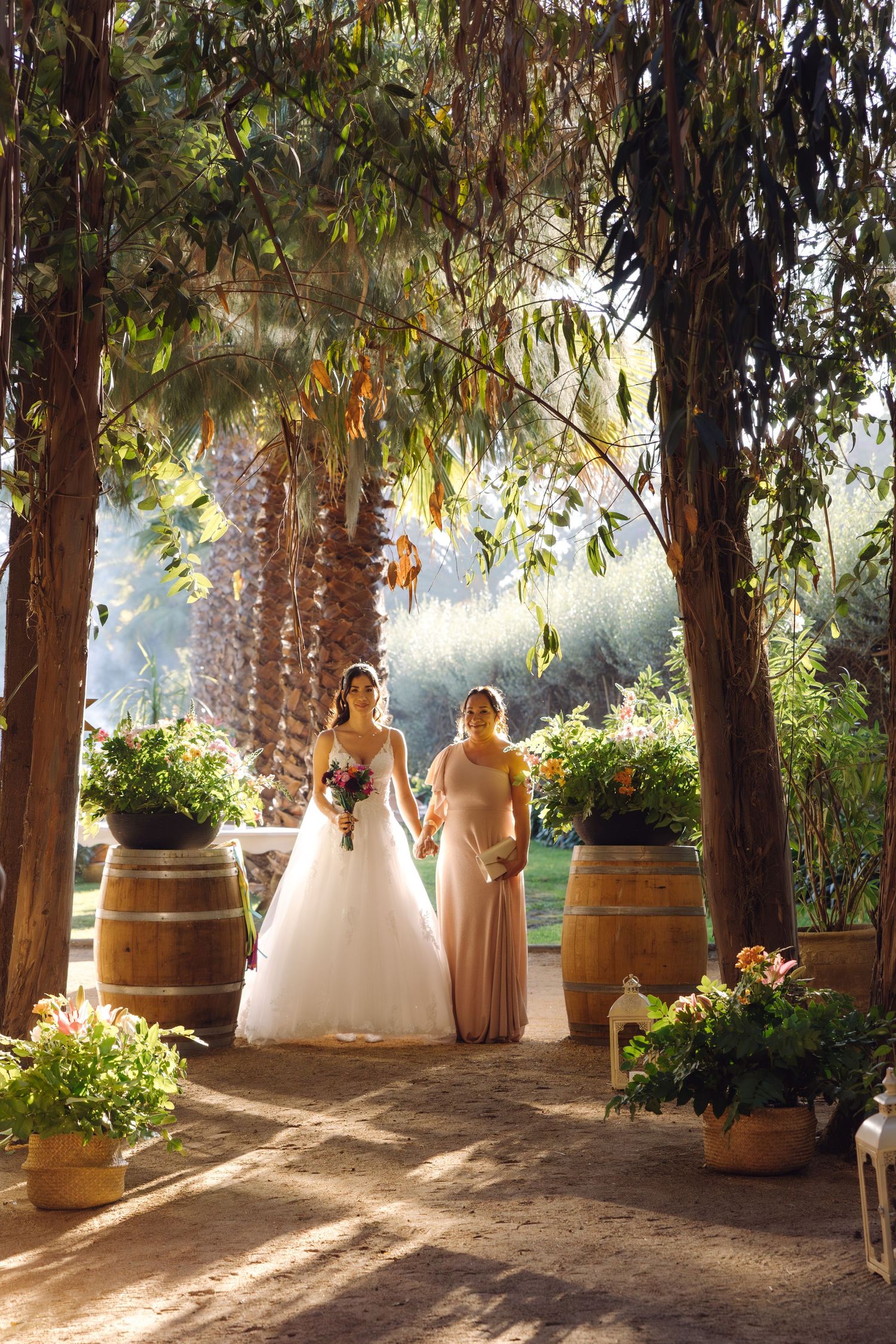 Bride in white gown walks down aisle with escort past wooden barrels in sunlit garden wedding.