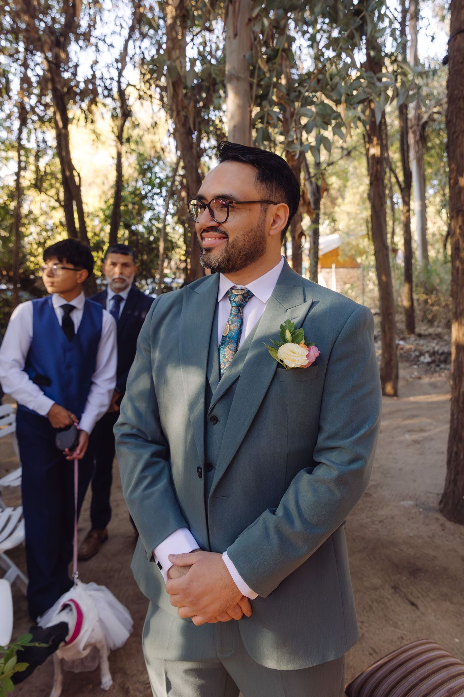 Smiling groom in gray suit with floral boutonniere awaits bride at outdoor woodland wedding.