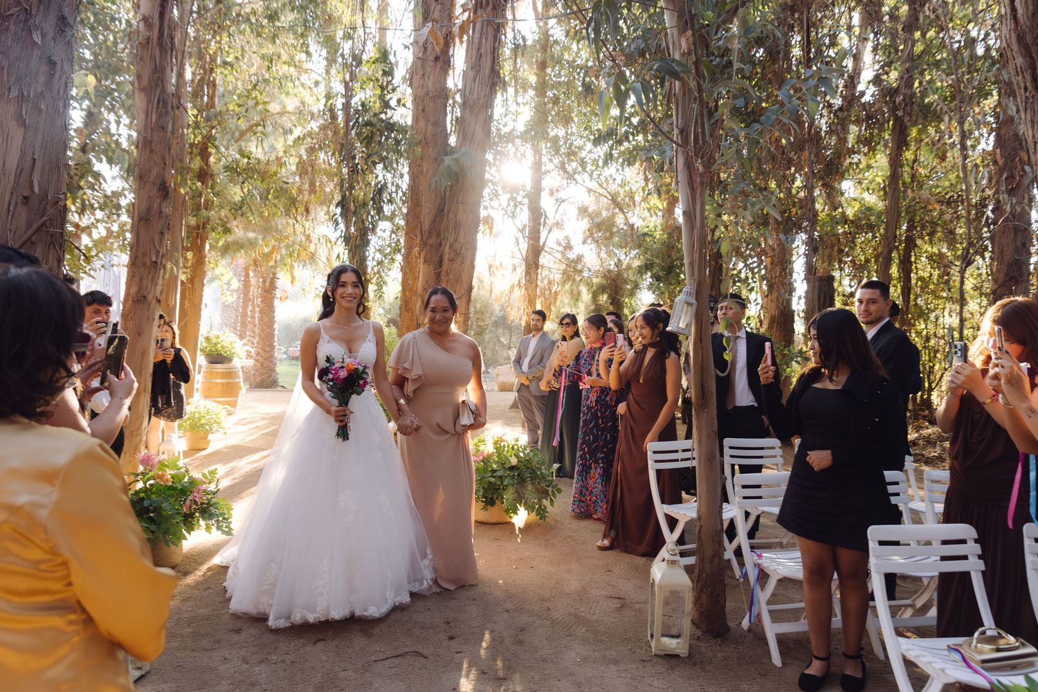 Bride in white gown walks down outdoor aisle with bridesmaid past seated wedding guests.