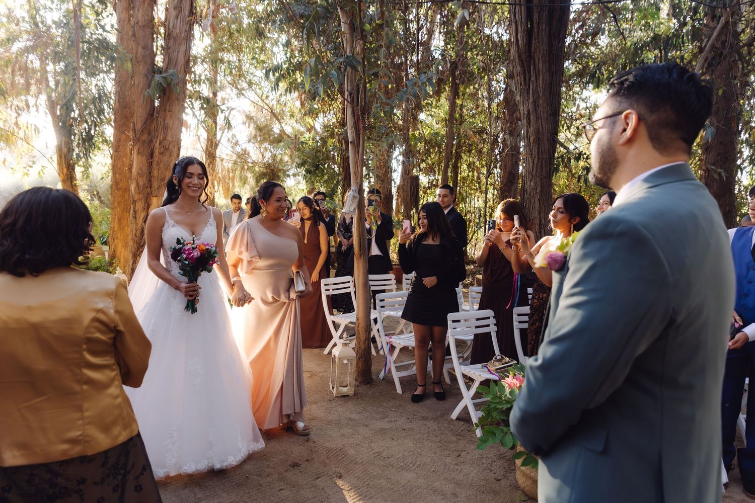 Bride in white gown approaches smiling groom at outdoor woodland wedding ceremony aisle.