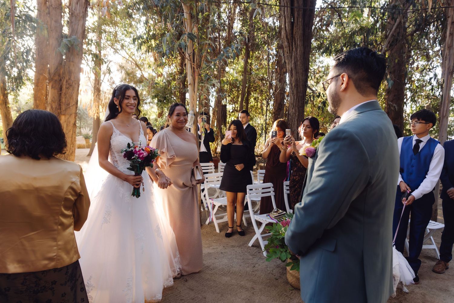 Bride in white dress walks down aisle toward groom at sunlit outdoor forest wedding ceremony.