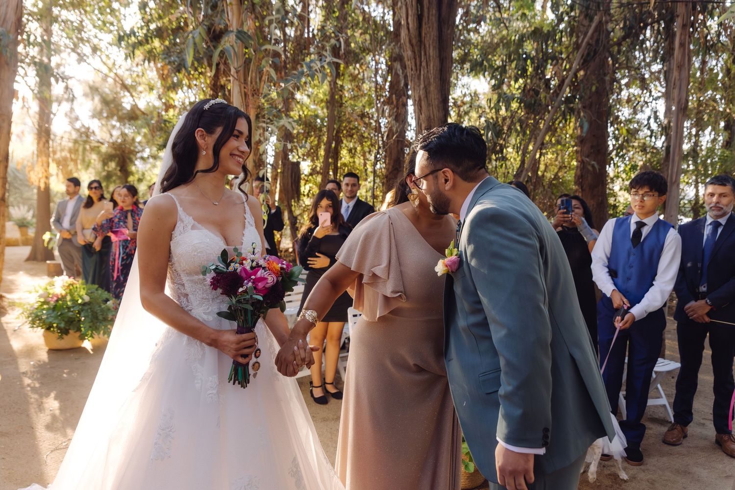 Groom kisses hand of smiling bride in white gown at outdoor woodland wedding ceremony.