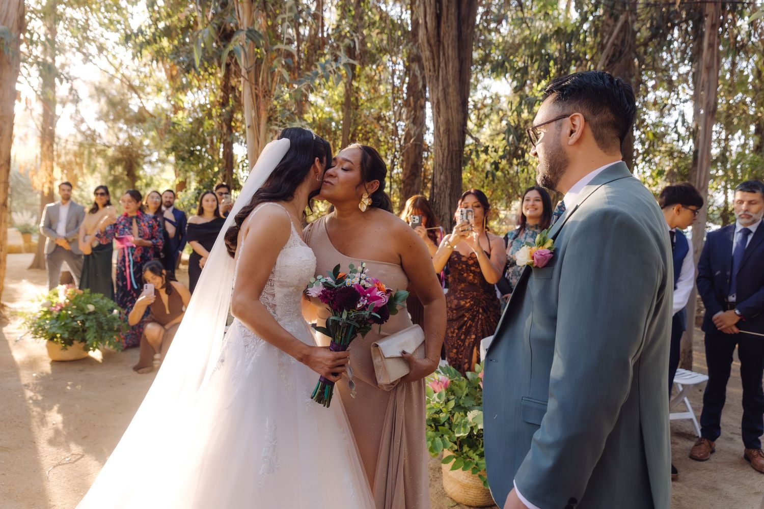 Bride and bridesmaid share an emotional kiss during outdoor wedding ceremony surrounded by tall trees and guests.