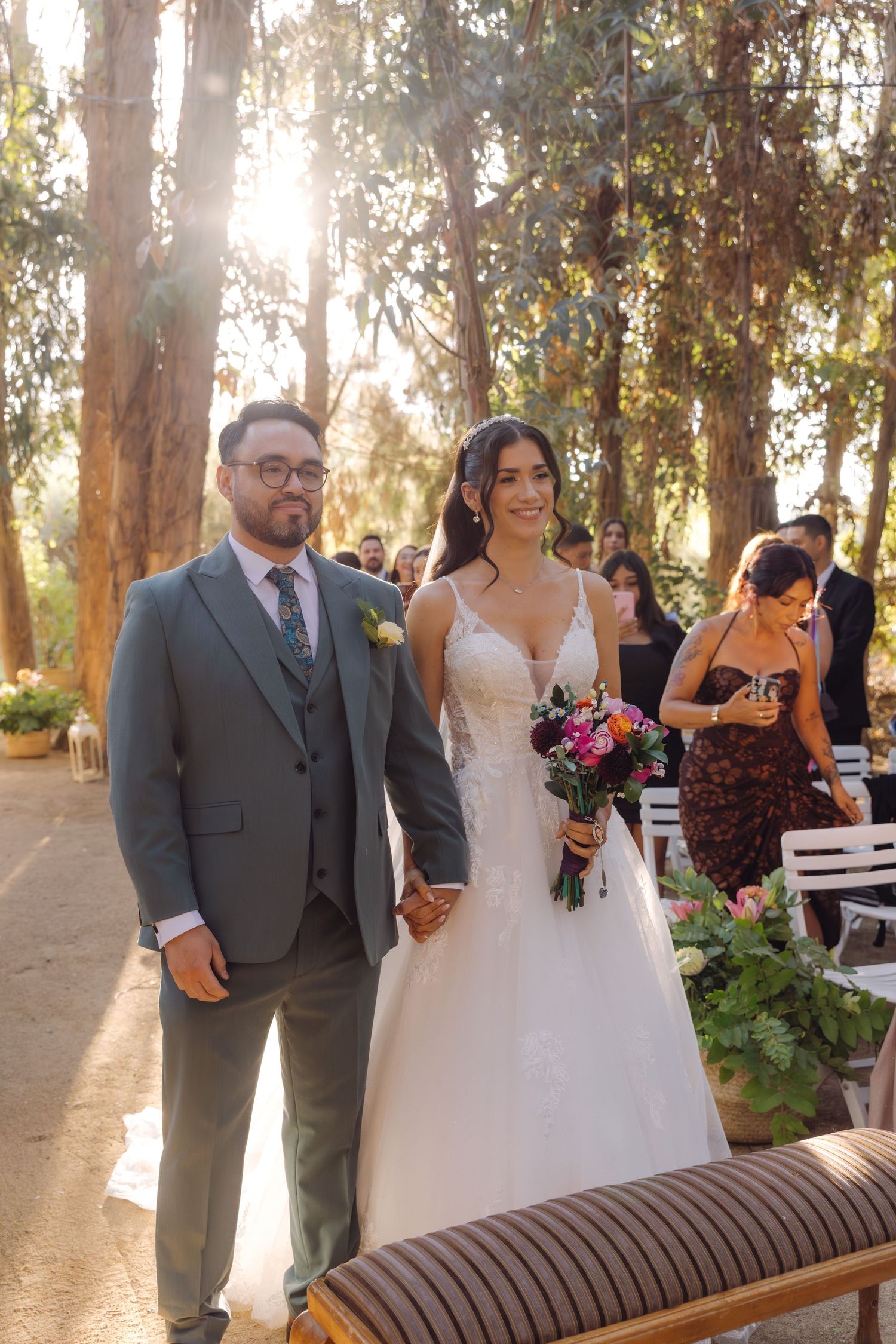 Smiling bride and groom walk down the aisle hand in hand at a sunlit outdoor forest wedding ceremony.