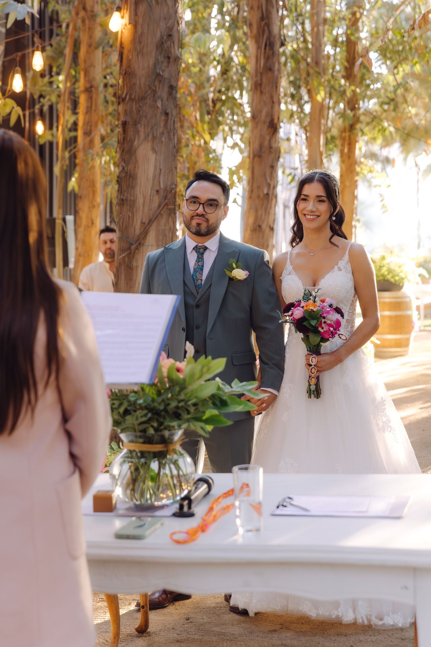 Bride and groom stand before officiant at elegant outdoor wedding ceremony altar surrounded by trees and warm light.
