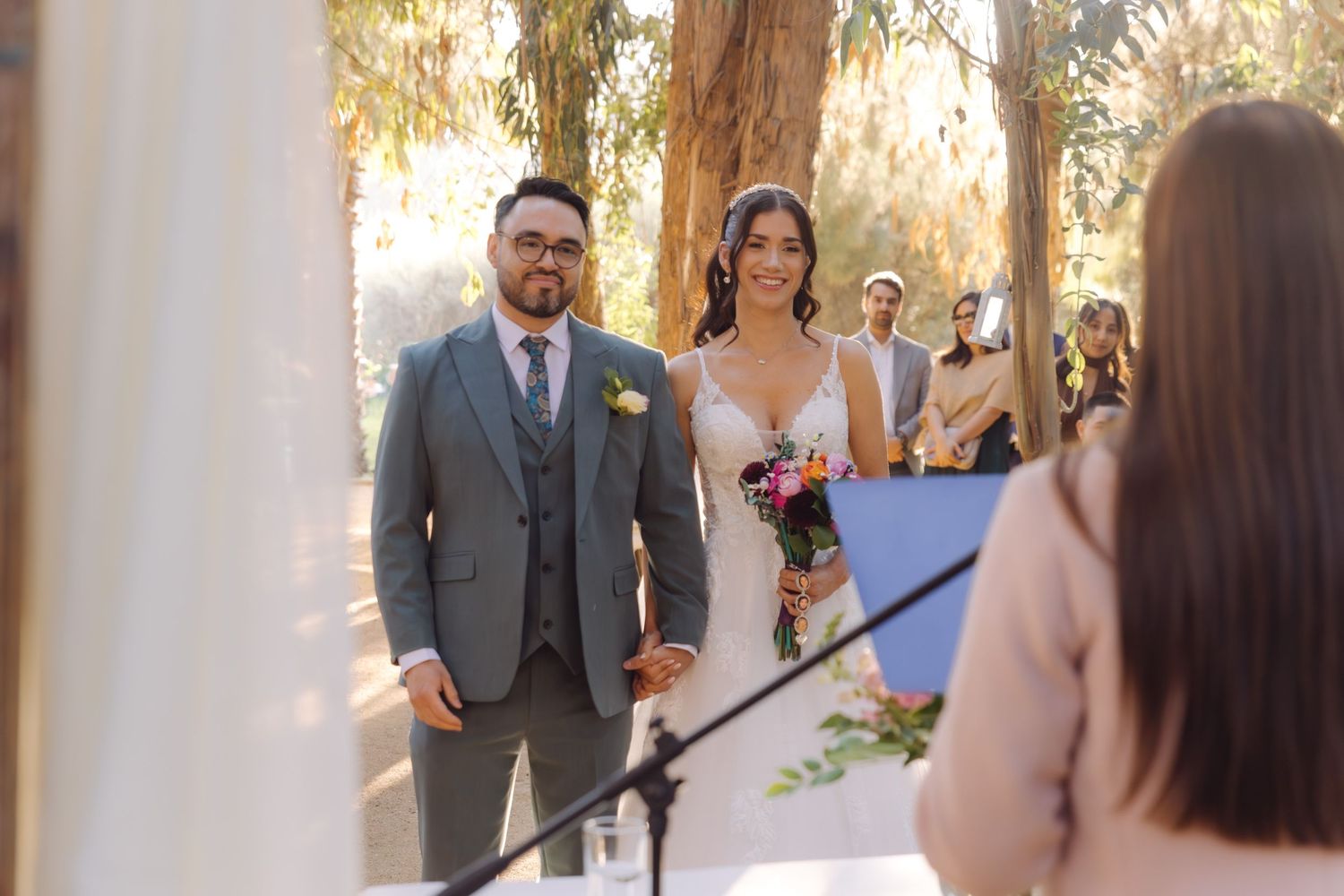 Groom in gray suit and bride holding colorful bouquet stand at outdoor woodland wedding ceremony altar.