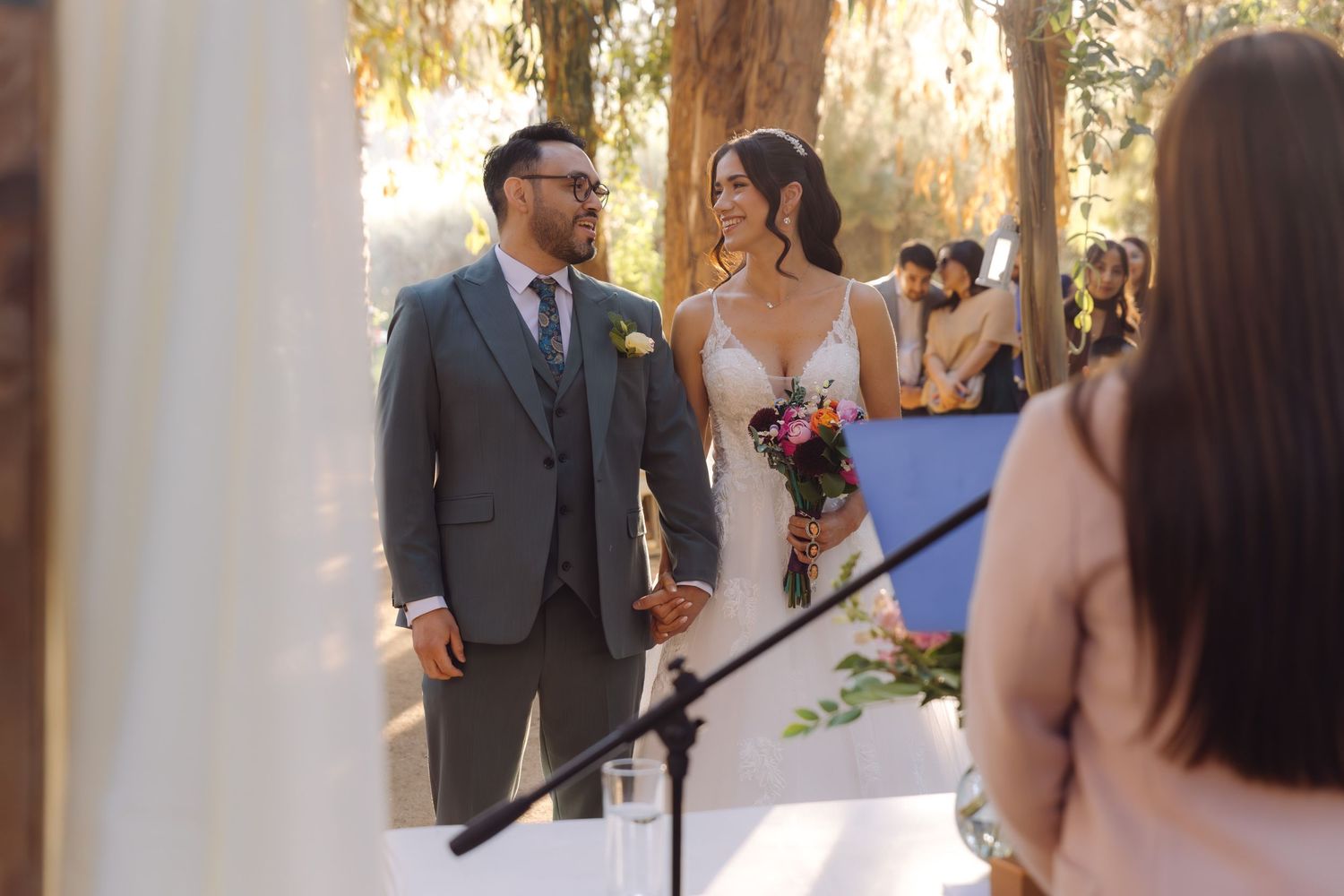 Bride and groom hold hands and smile at each other during a beautiful sunlit outdoor forest wedding ceremony.