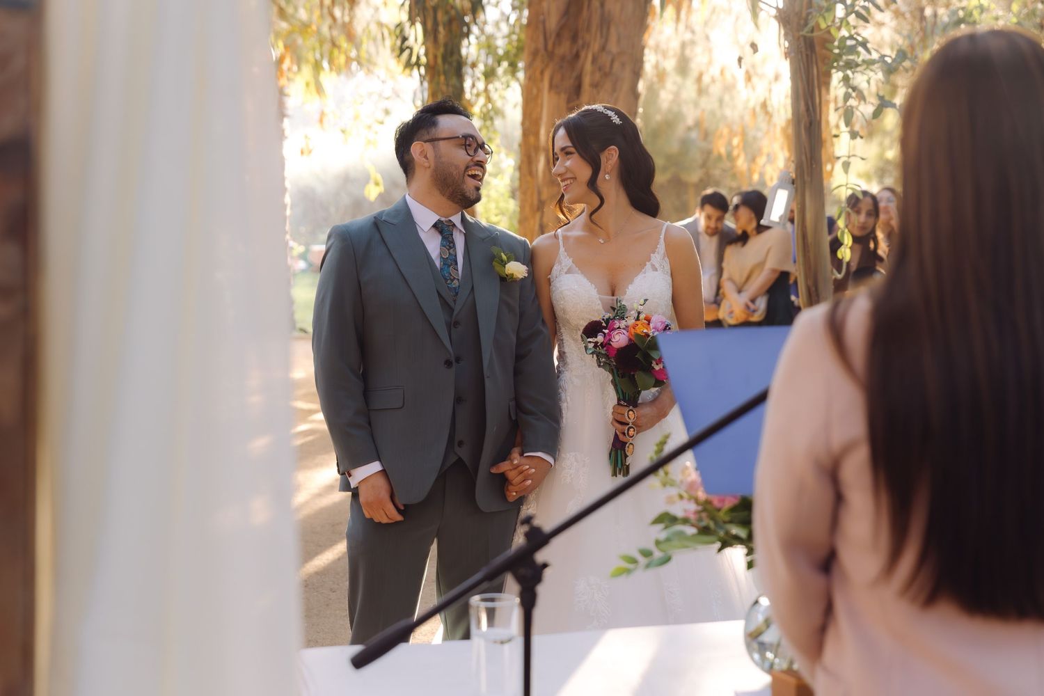 Groom smiles lovingly at bride during outdoor woodland wedding ceremony with warm golden sunlight filtering through trees.