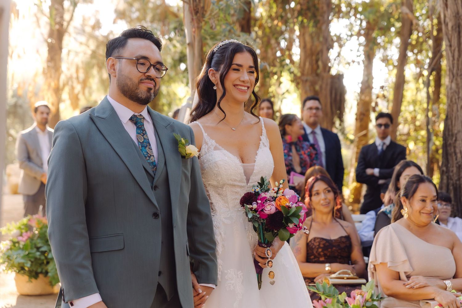 Smiling bride holding colorful bouquet and groom in gray suit walk together at sunny outdoor forest wedding.